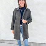 A woman stands outdoors against a white stucco wall wearing a long brown and black plaid coat with faux leather collar over a black turtleneck, blue jeans, and brown mesh Mary Jane flats.