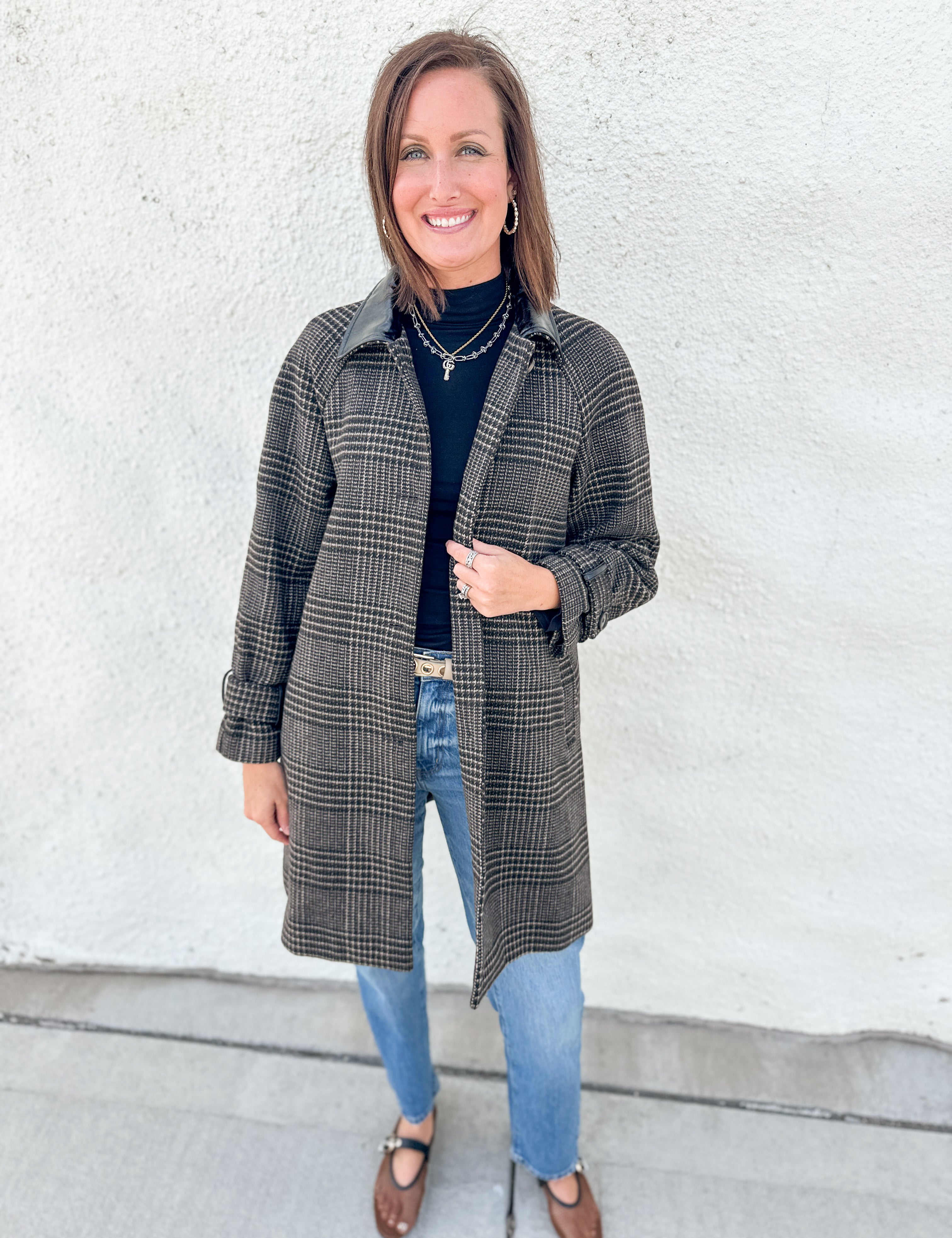 A woman stands outdoors against a white stucco wall wearing a long brown and black plaid coat with faux leather collar over a black turtleneck, blue jeans, and brown mesh Mary Jane flats.
