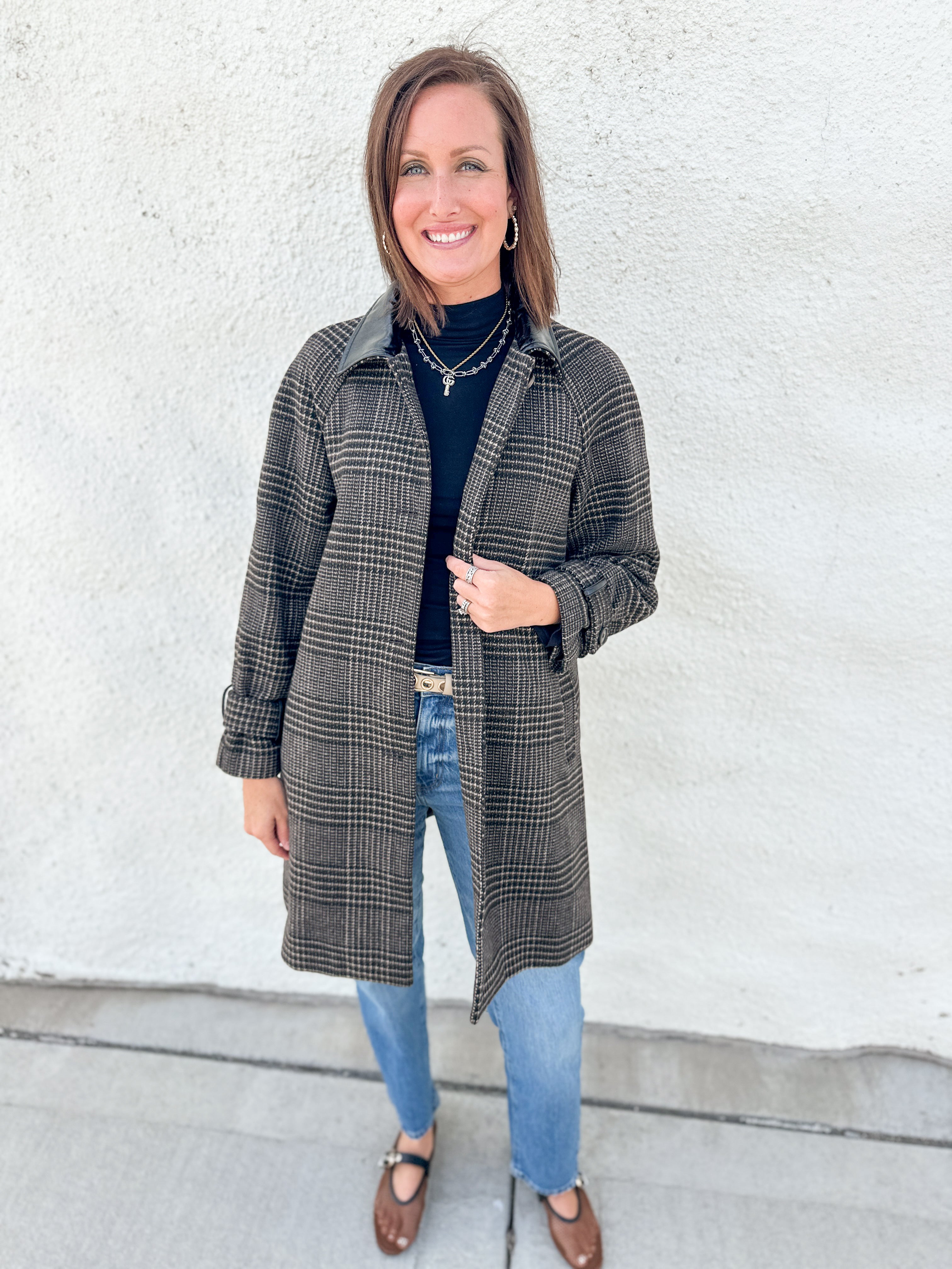 A woman stands outdoors against a white stucco wall wearing a long brown and black plaid coat with faux leather collar over a black turtleneck, blue jeans, and brown mesh Mary Jane flats.