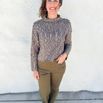 A woman stands outdoors against a white stucco wall wearing a textured, cable-knit mock neck sweater in a heathered brown and gray colorway, paired with olive green pants and dark loafers.