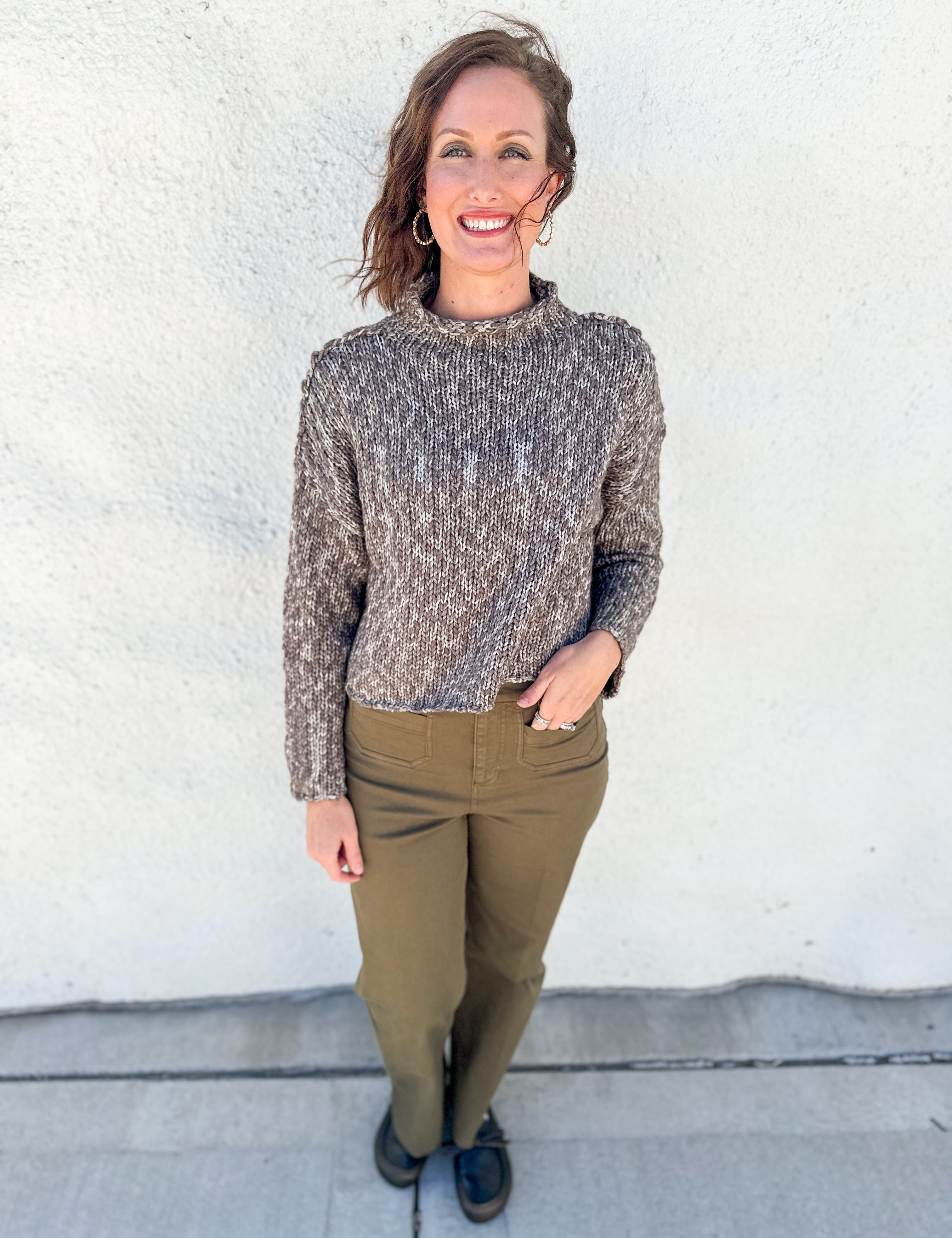 A woman stands outdoors against a white stucco wall wearing a textured, cable-knit mock neck sweater in a heathered brown and gray colorway, paired with olive green pants and dark loafers.