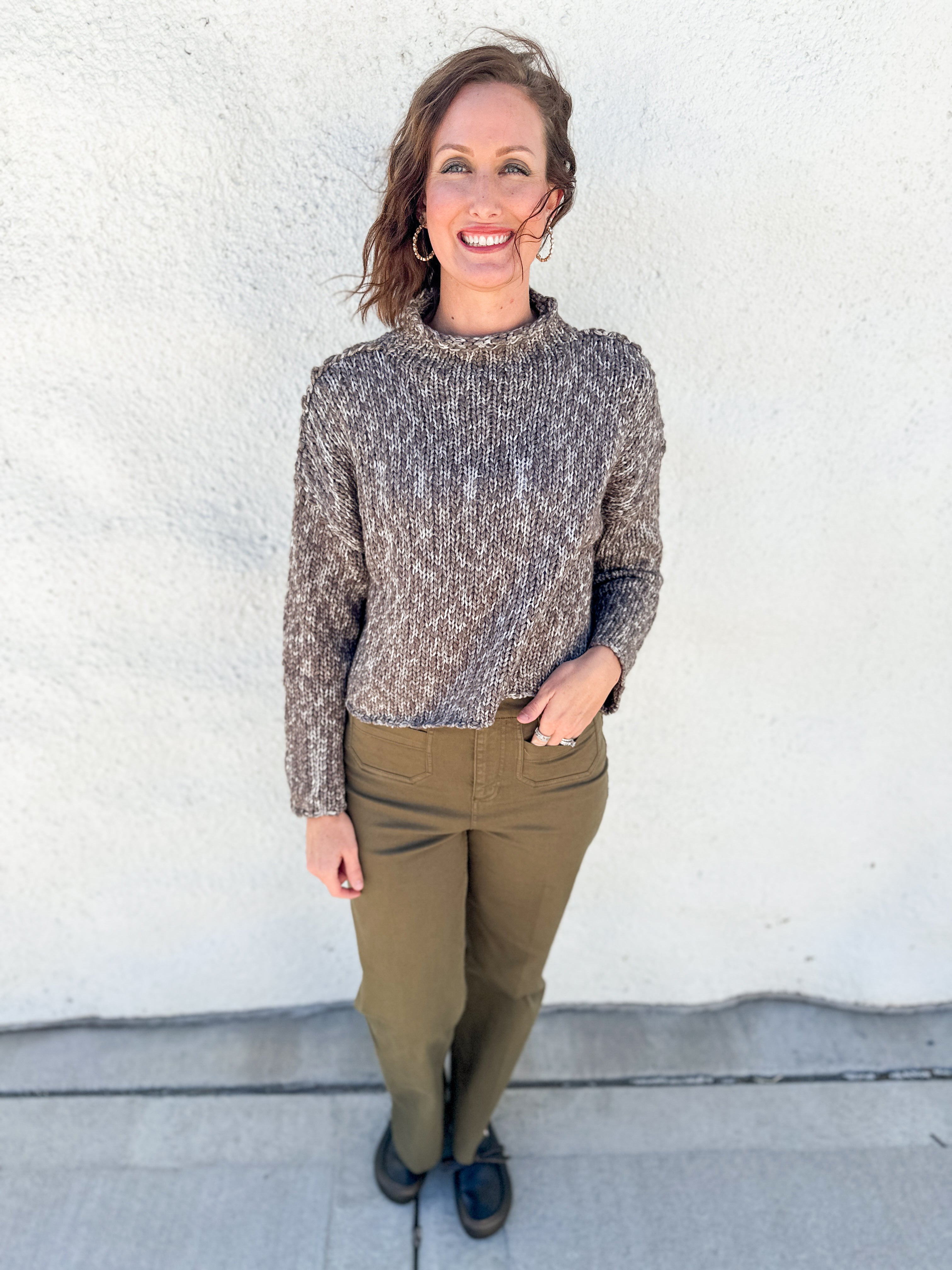 A woman stands outdoors against a white stucco wall wearing a textured, cable-knit mock neck sweater in a heathered brown and gray colorway, paired with olive green pants and dark loafers.