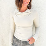 A woman wearing a fitted long-sleeve cowl neck top in cream and gray jeans, standing against a textured white wall.
