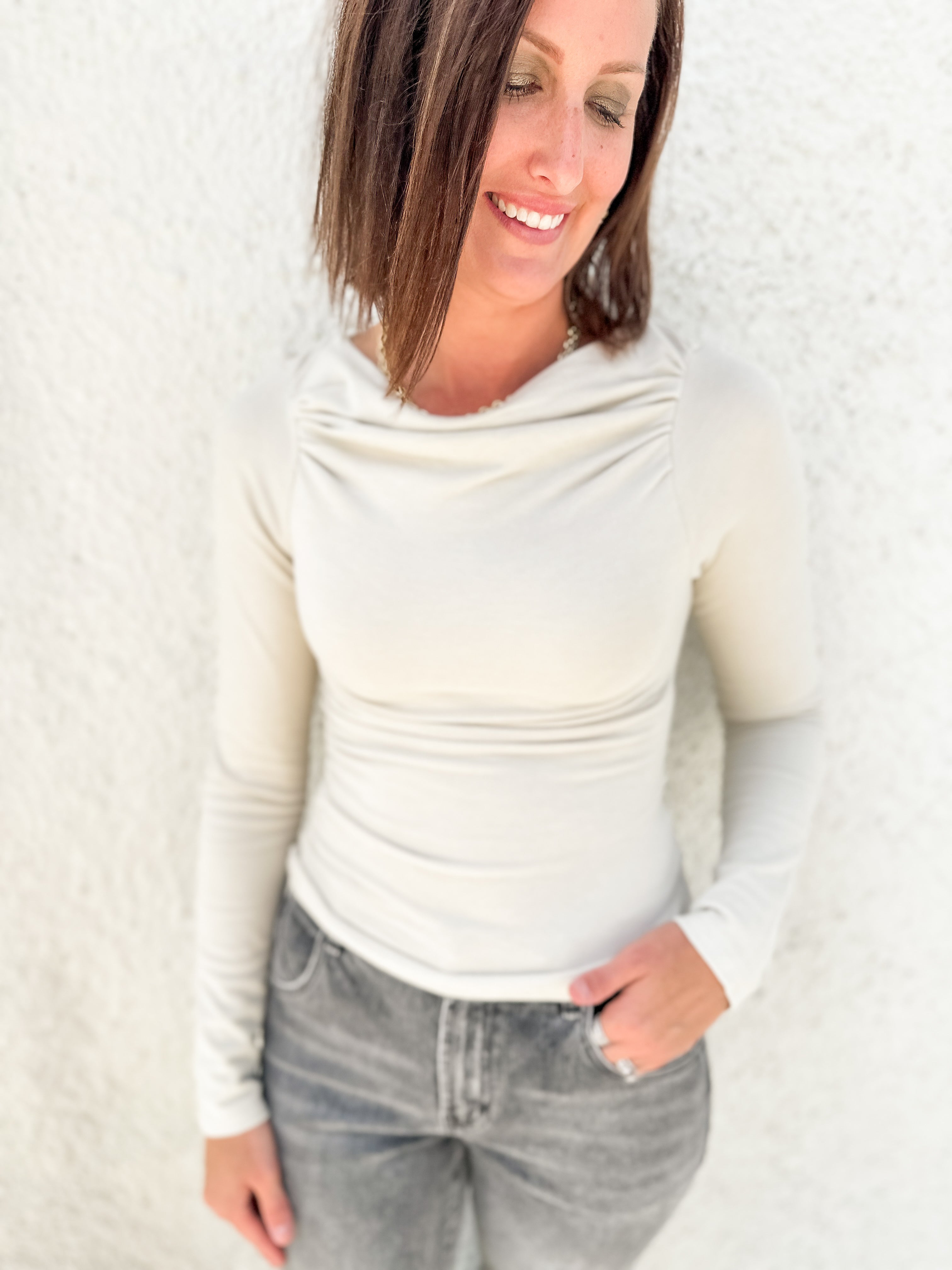 A woman wearing a fitted long-sleeve cowl neck top in cream and gray jeans, standing against a textured white wall.