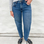 A person wearing blue barrel leg jeans, a light gray ribbed sweater, and distressed black loafer clog slip-on shoes, standing against a white textured wall.
