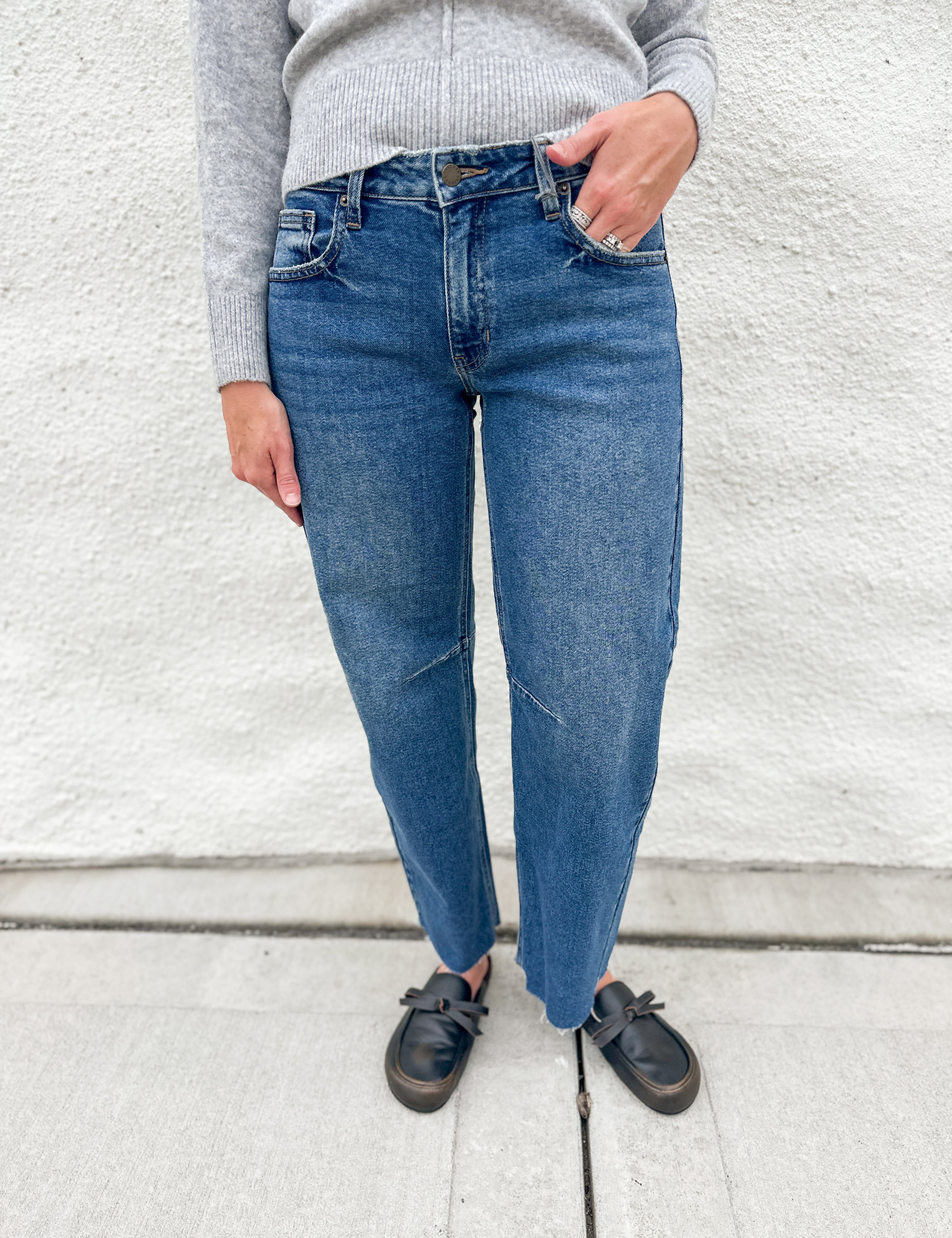 A person wearing blue barrel leg jeans, a light gray ribbed sweater, and distressed black loafer clog slip-on shoes, standing against a white textured wall.