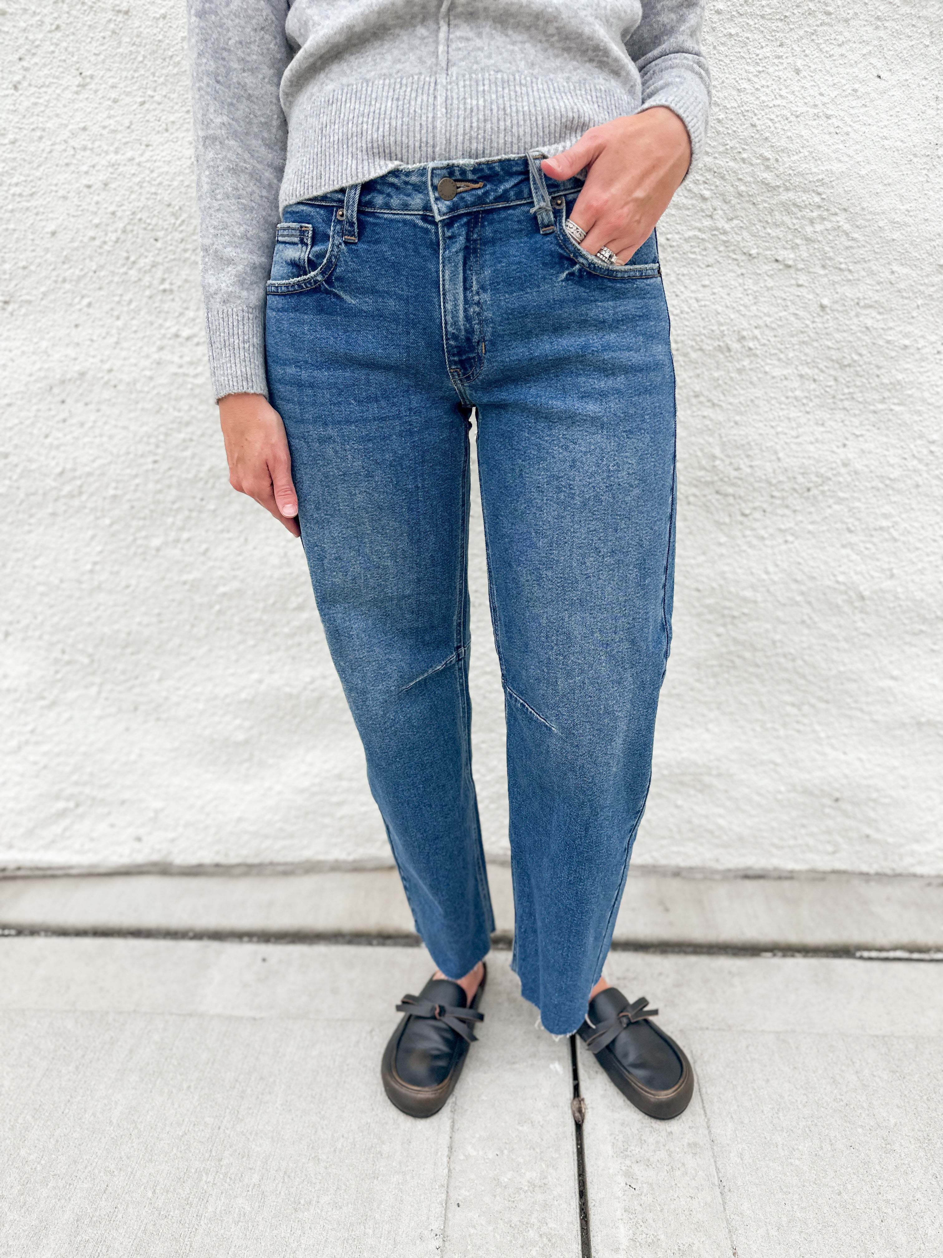A person wearing blue barrel leg jeans, a light gray ribbed sweater, and distressed black loafer clog slip-on shoes, standing against a white textured wall.