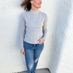 A woman wearing a light gray mock neck knit sweater and blue jeans, standing against a white textured stucco wall.