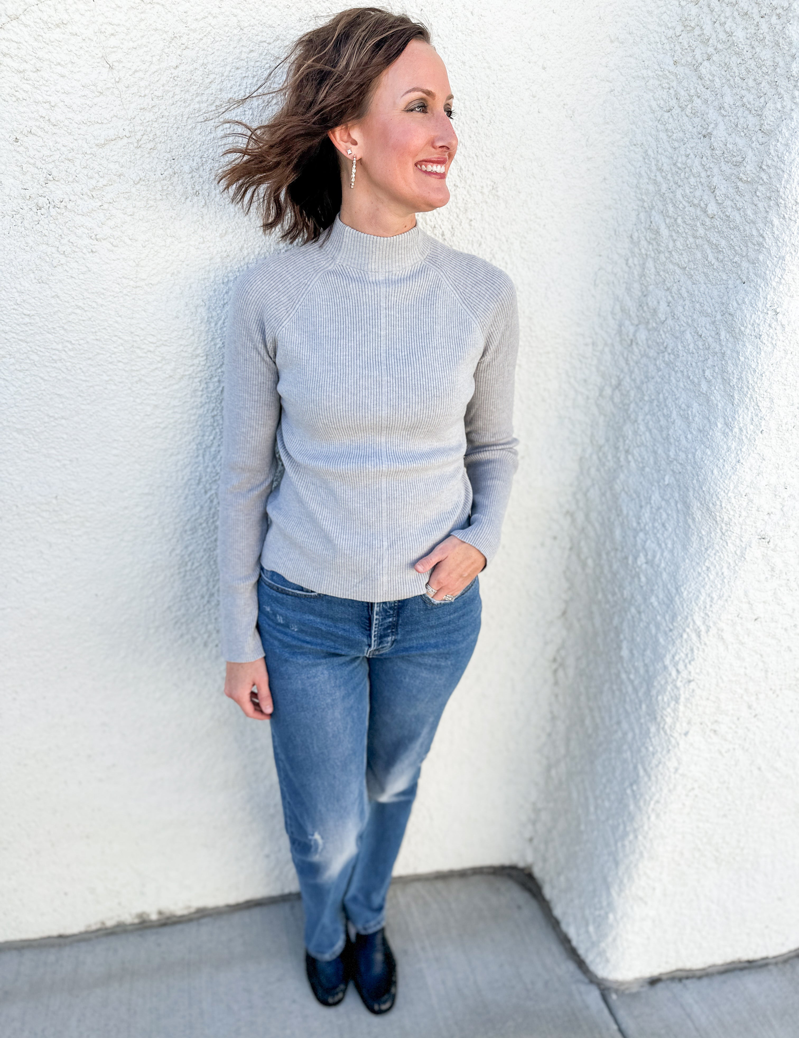 A woman wearing a light gray mock neck knit sweater and blue jeans, standing against a white textured stucco wall.