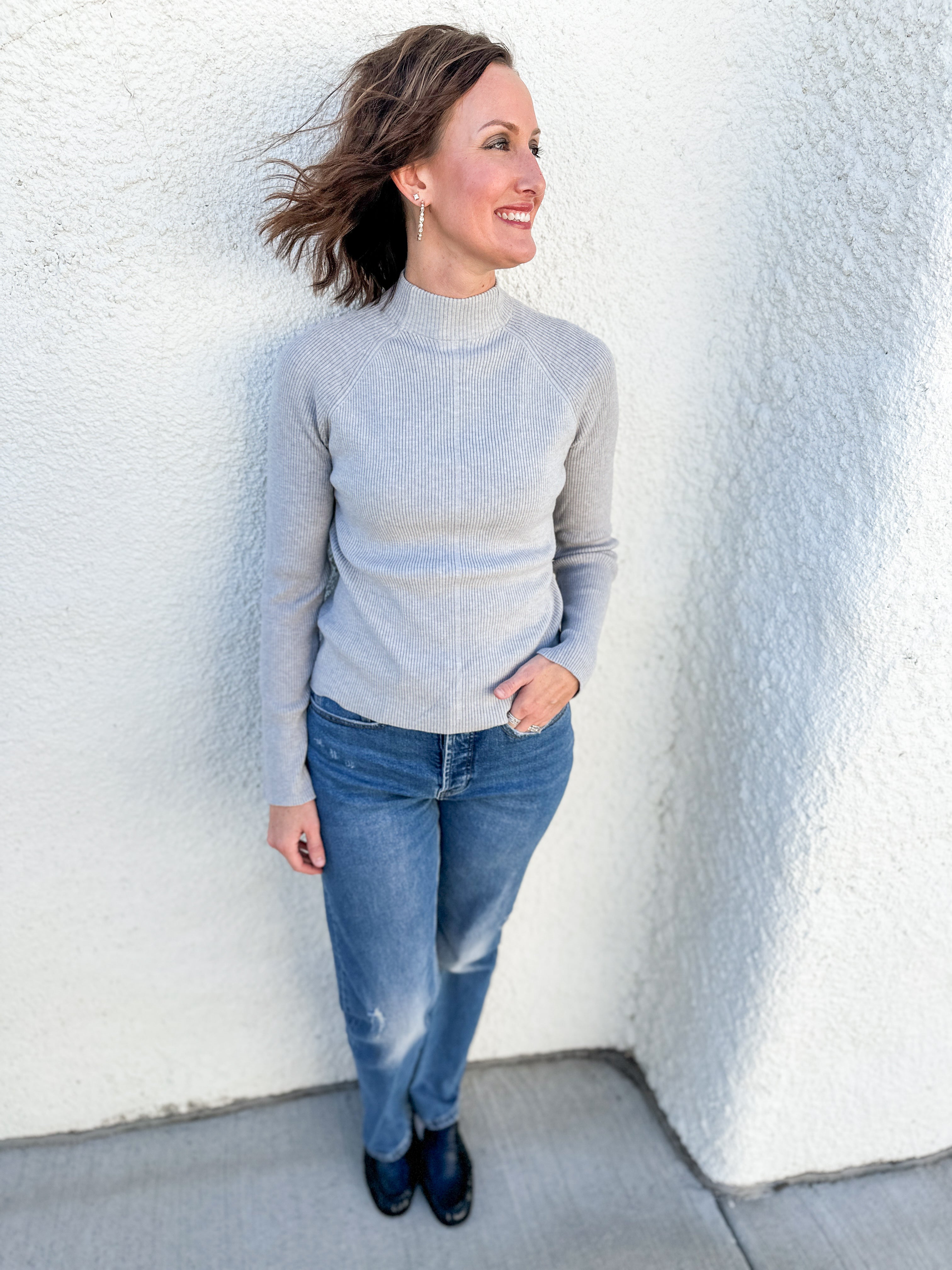 A woman wearing a light gray mock neck knit sweater and blue jeans, standing against a white textured stucco wall.