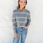A woman wearing a gray and white striped knit sweater with blue jeans, standing against a white textured wall.