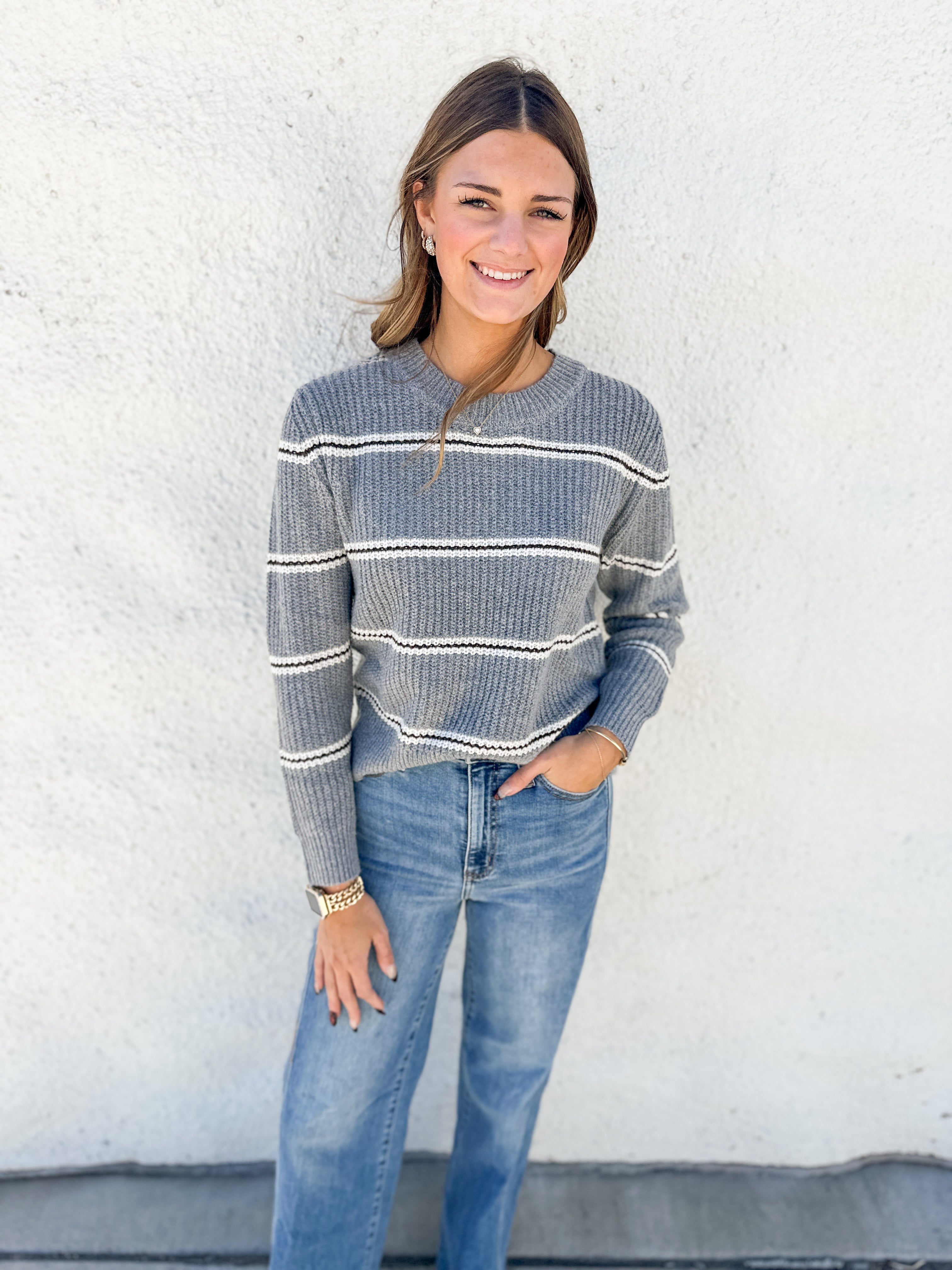 A woman wearing a gray and white striped knit sweater with blue jeans, standing against a white textured wall.