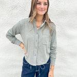 A woman modeling a light green heathered long-sleeve button-up shirt with a chest pocket, paired with dark blue jeans, standing against a white textured wall.