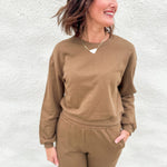 A woman wearing a matching brown sweatshirt and jogger pants set, standing against a white textured wall.