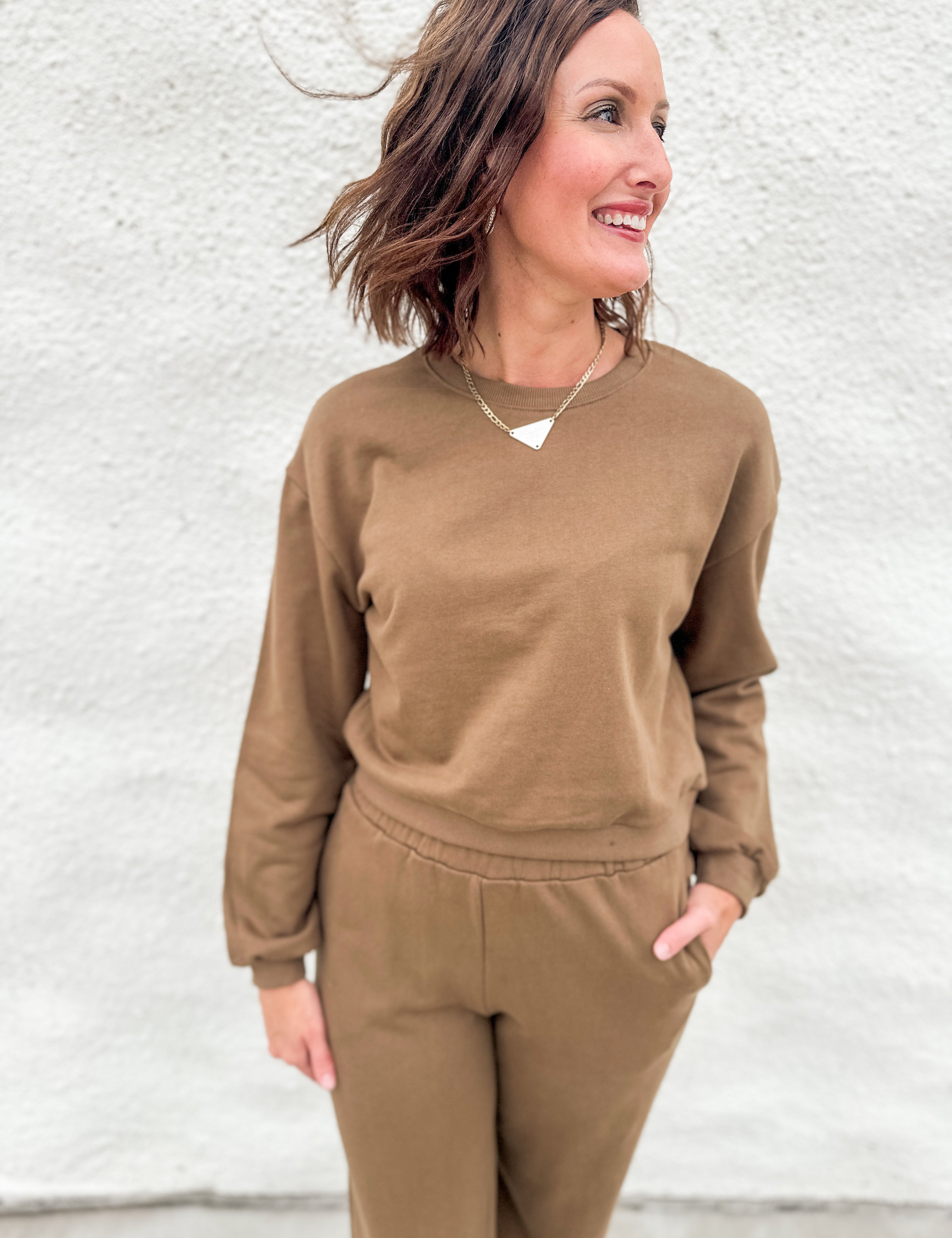 A woman wearing a matching brown sweatshirt and jogger pants set, standing against a white textured wall.