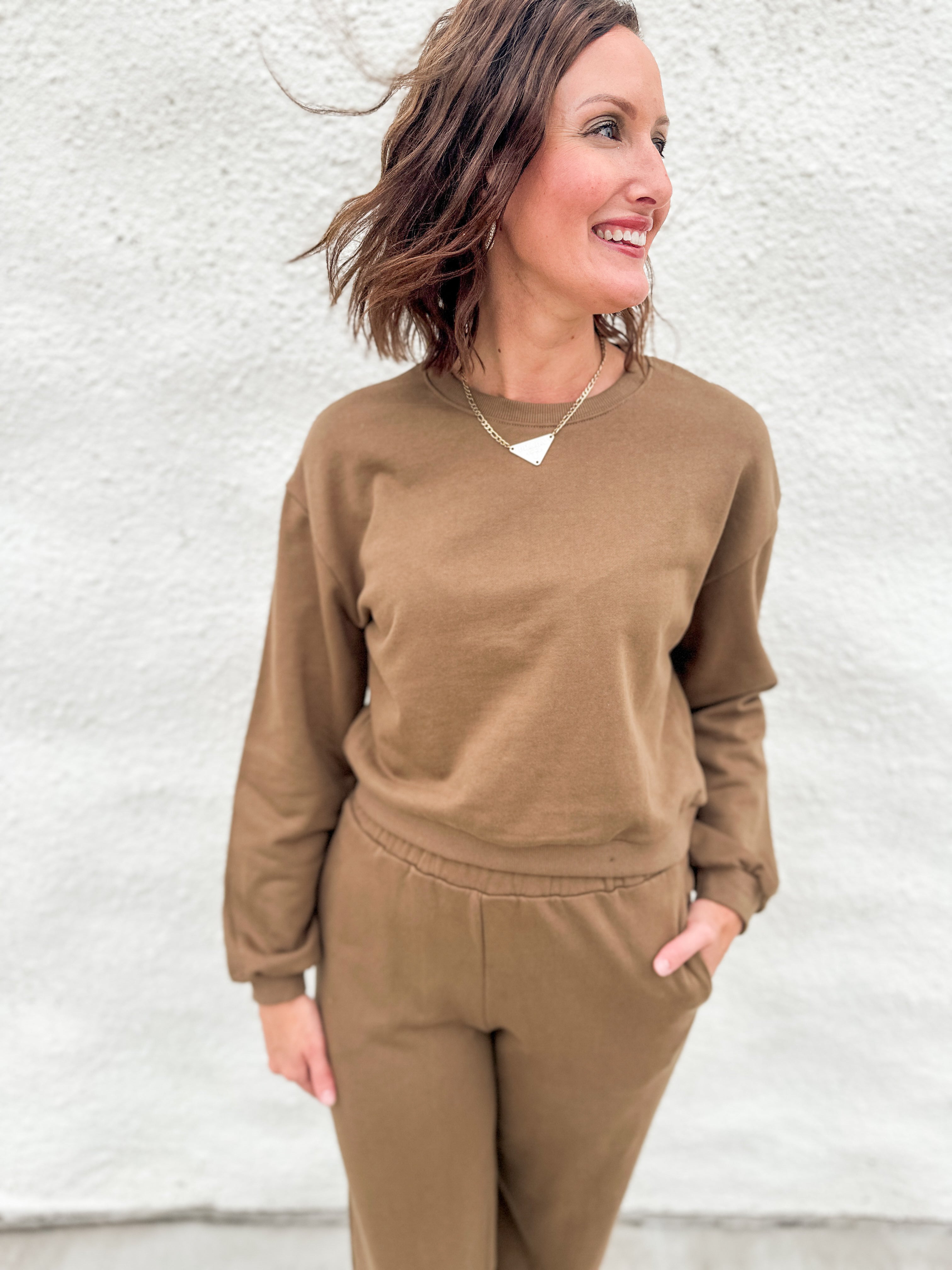 A woman wearing a matching brown sweatshirt and jogger pants set, standing against a white textured wall.