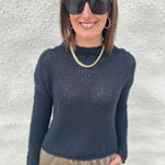 A woman wearing a black opal knit mock neck sweater from Free People, black sunglasses, a gold chain necklace, and khaki drawstring pants, standing against a textured white wall.