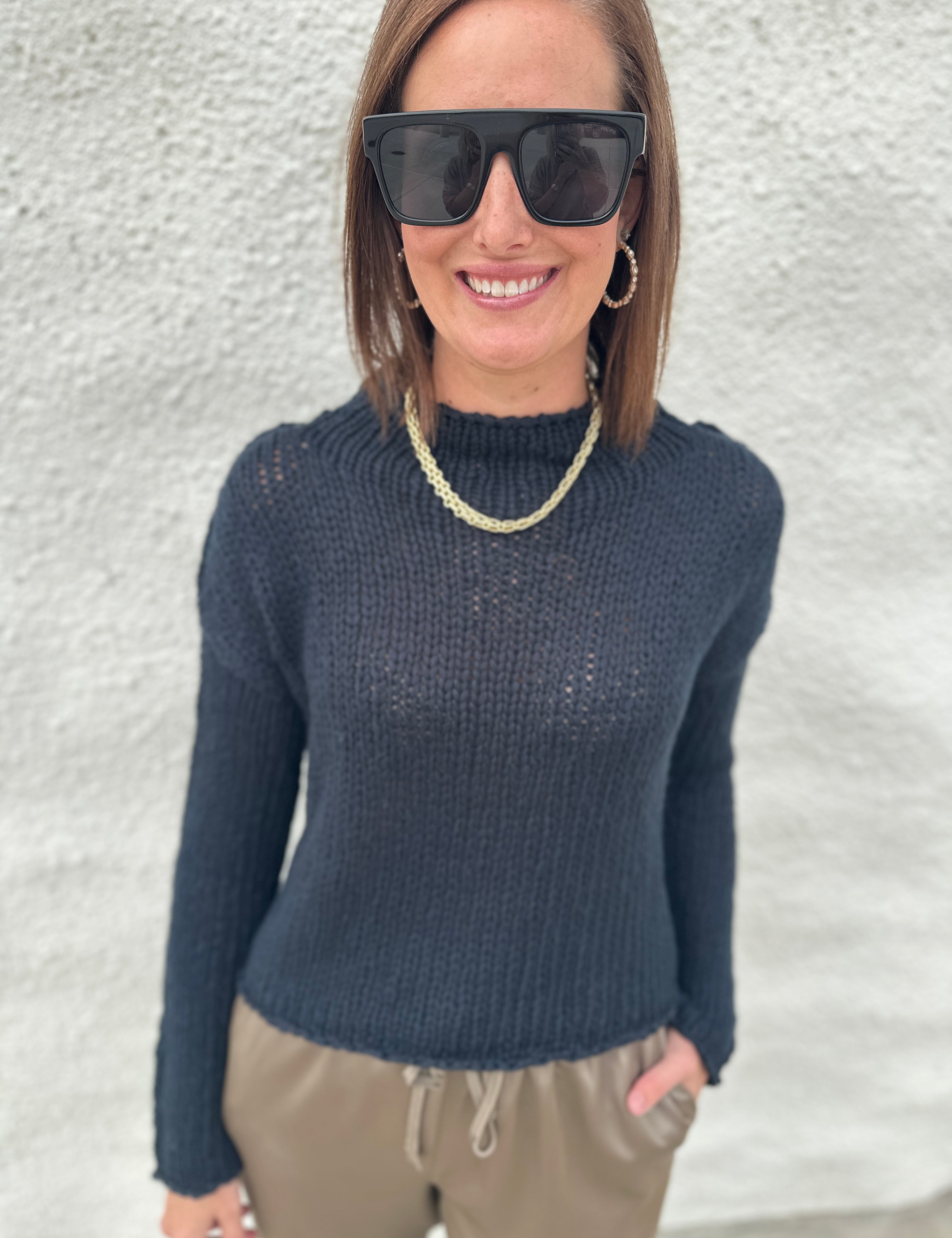 A woman wearing a black opal knit mock neck sweater from Free People, black sunglasses, a gold chain necklace, and khaki drawstring pants, standing against a textured white wall.