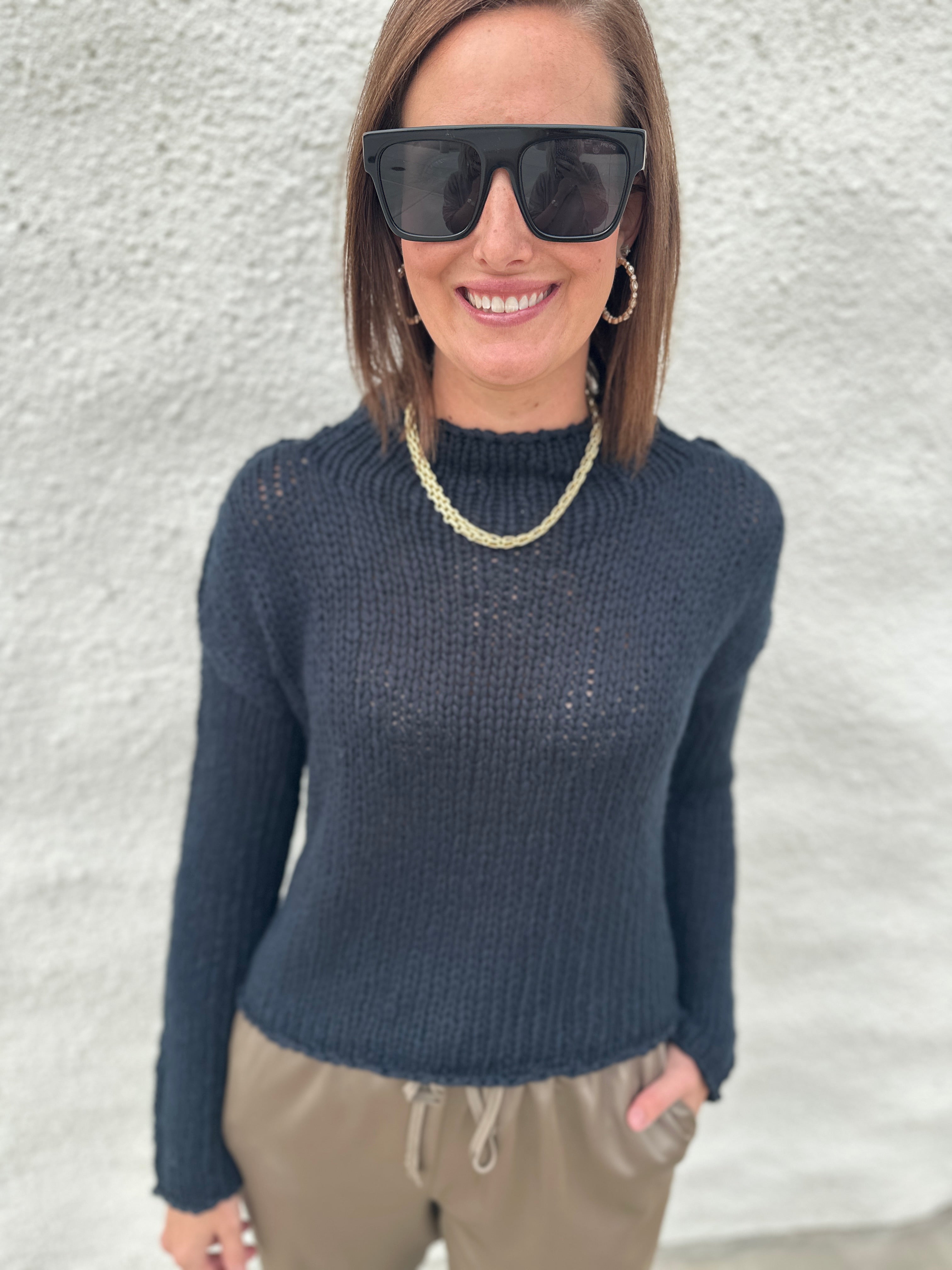 A woman wearing a black opal knit mock neck sweater from Free People, black sunglasses, a gold chain necklace, and khaki drawstring pants, standing against a textured white wall.