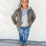 A woman stands against a white stucco wall wearing an olive green quilted puffer jacket over a light gray sweater with white polka dots, blue jeans, and brown snake skin clogs.