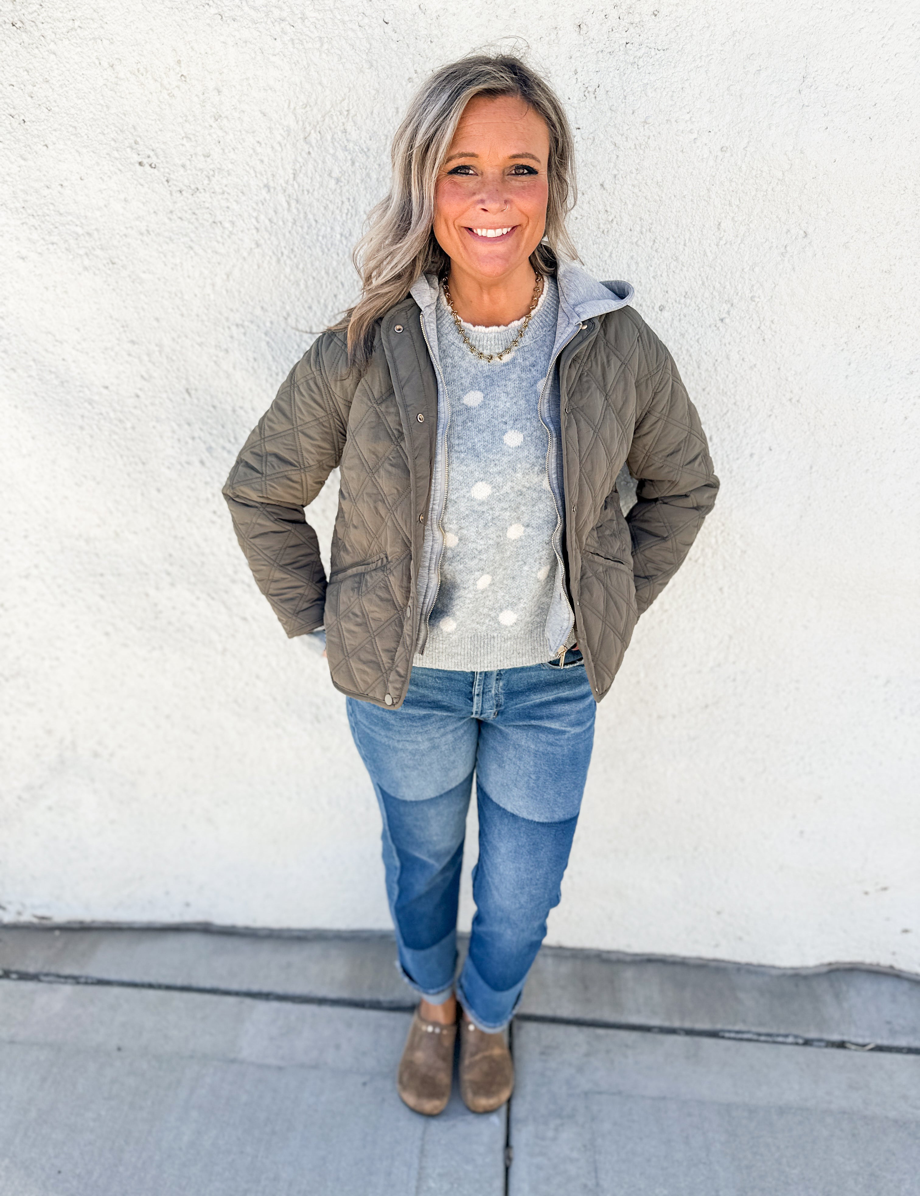 A woman stands against a white stucco wall wearing an olive green quilted puffer jacket over a light gray sweater with white polka dots, blue jeans, and brown snake skin clogs.