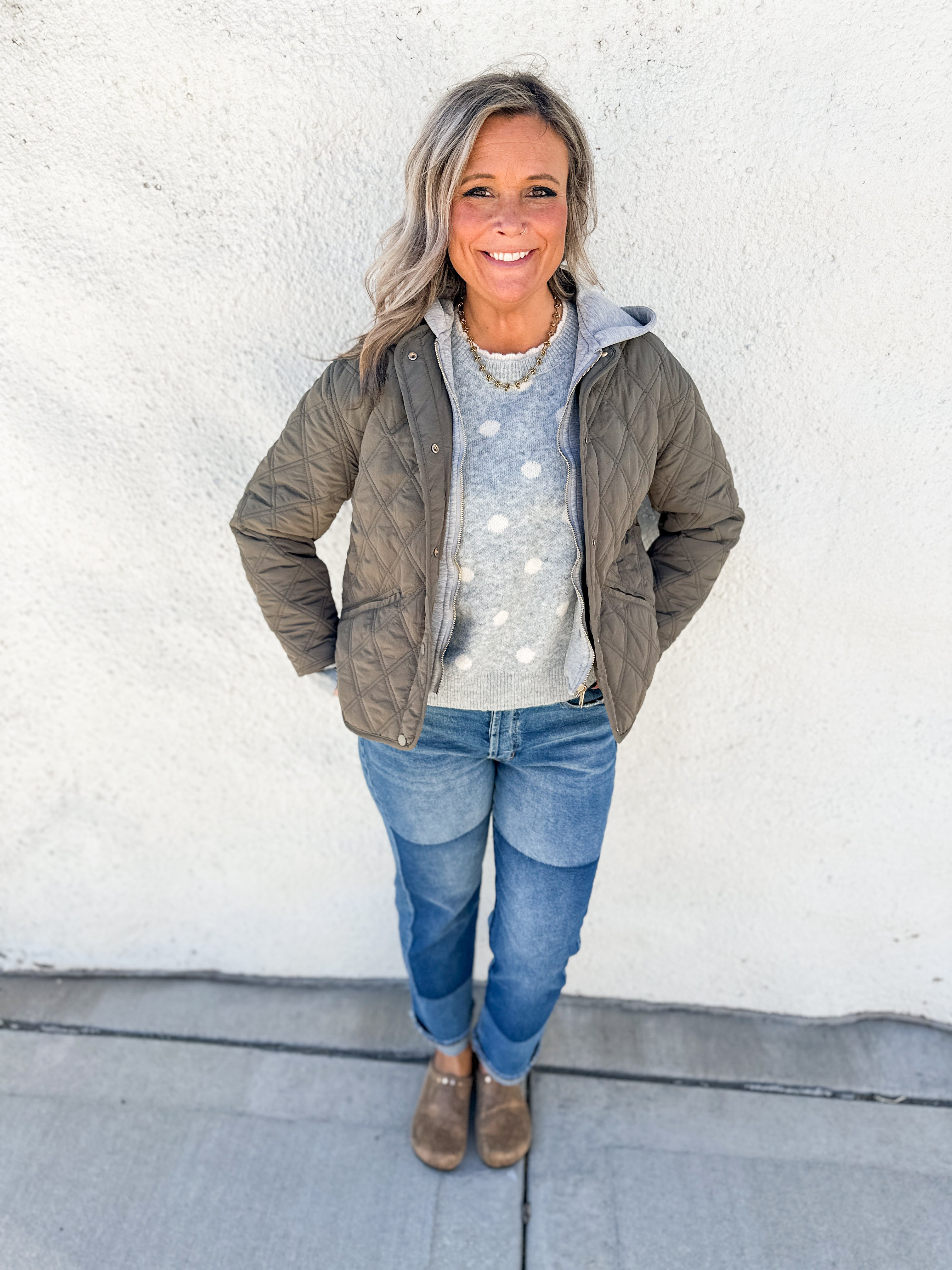 A woman stands against a white stucco wall wearing an olive green quilted puffer jacket over a light gray sweater with white polka dots, blue jeans, and brown snake skin clogs.