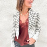 A woman wearing a black and white gingham blazer with white lace trim over a rust-brown lace camisole, paired with dark gray pants and black sunglasses, posing against a textured white wall.