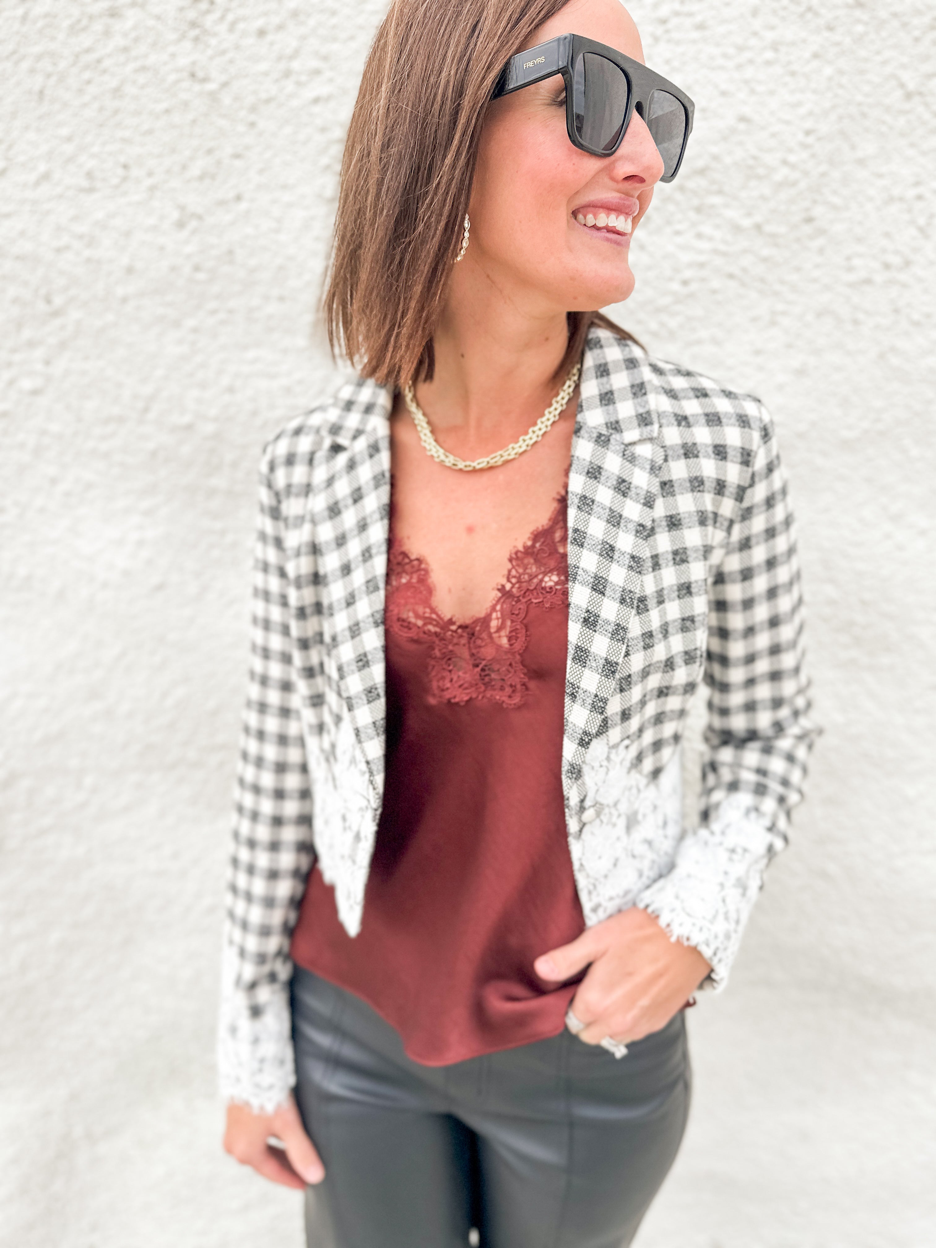 A woman wearing a black and white gingham blazer with white lace trim over a rust-brown lace camisole, paired with dark gray pants and black sunglasses, posing against a textured white wall.