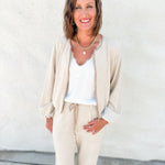 A woman smiling and posing in a beige bubble hem sweater bomber jacket with suede accents and matching drawstring pants over a white top, standing against a textured white stucco wall.