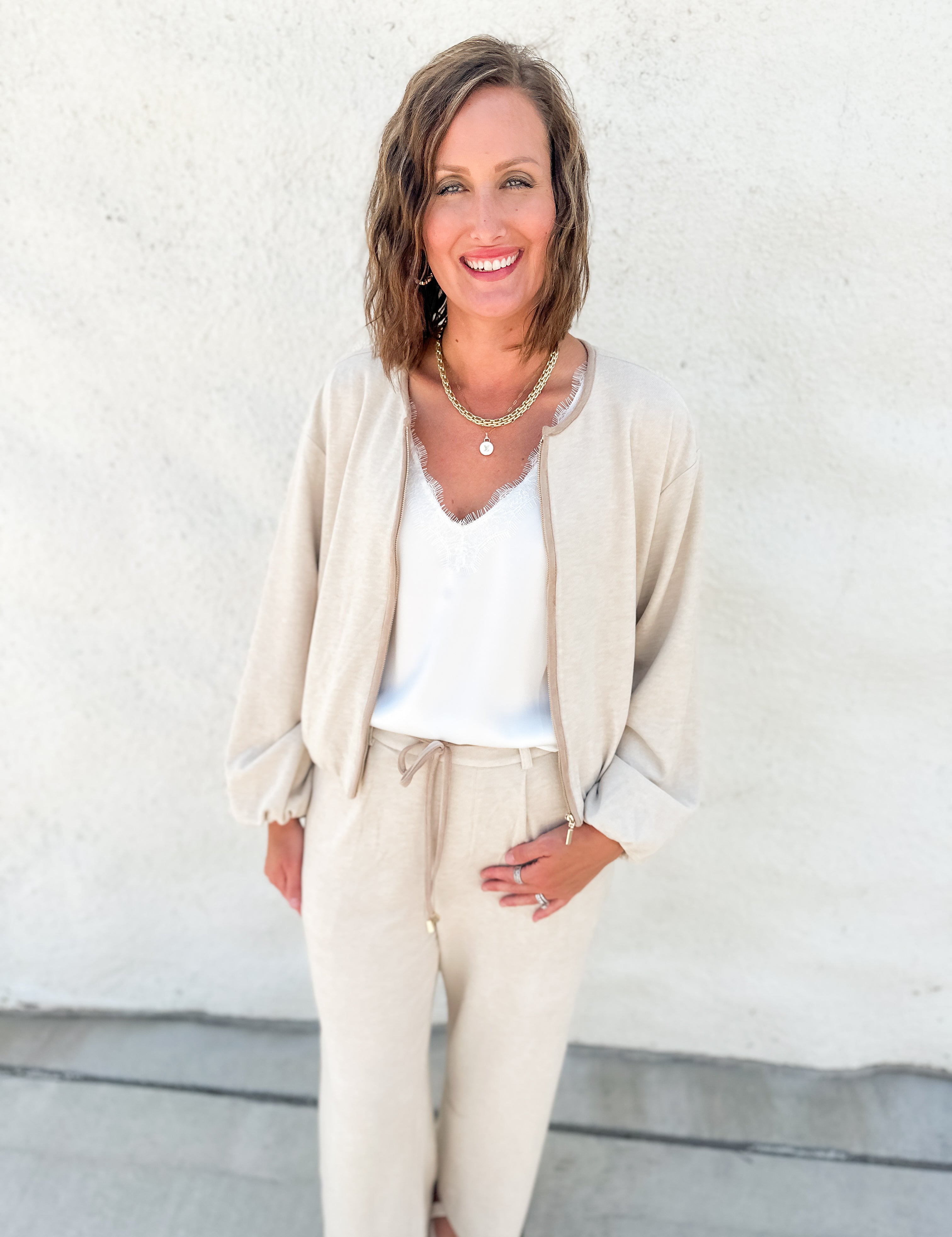 A woman smiling and posing in a beige bubble hem sweater bomber jacket with suede accents and matching drawstring pants over a white top, standing against a textured white stucco wall.