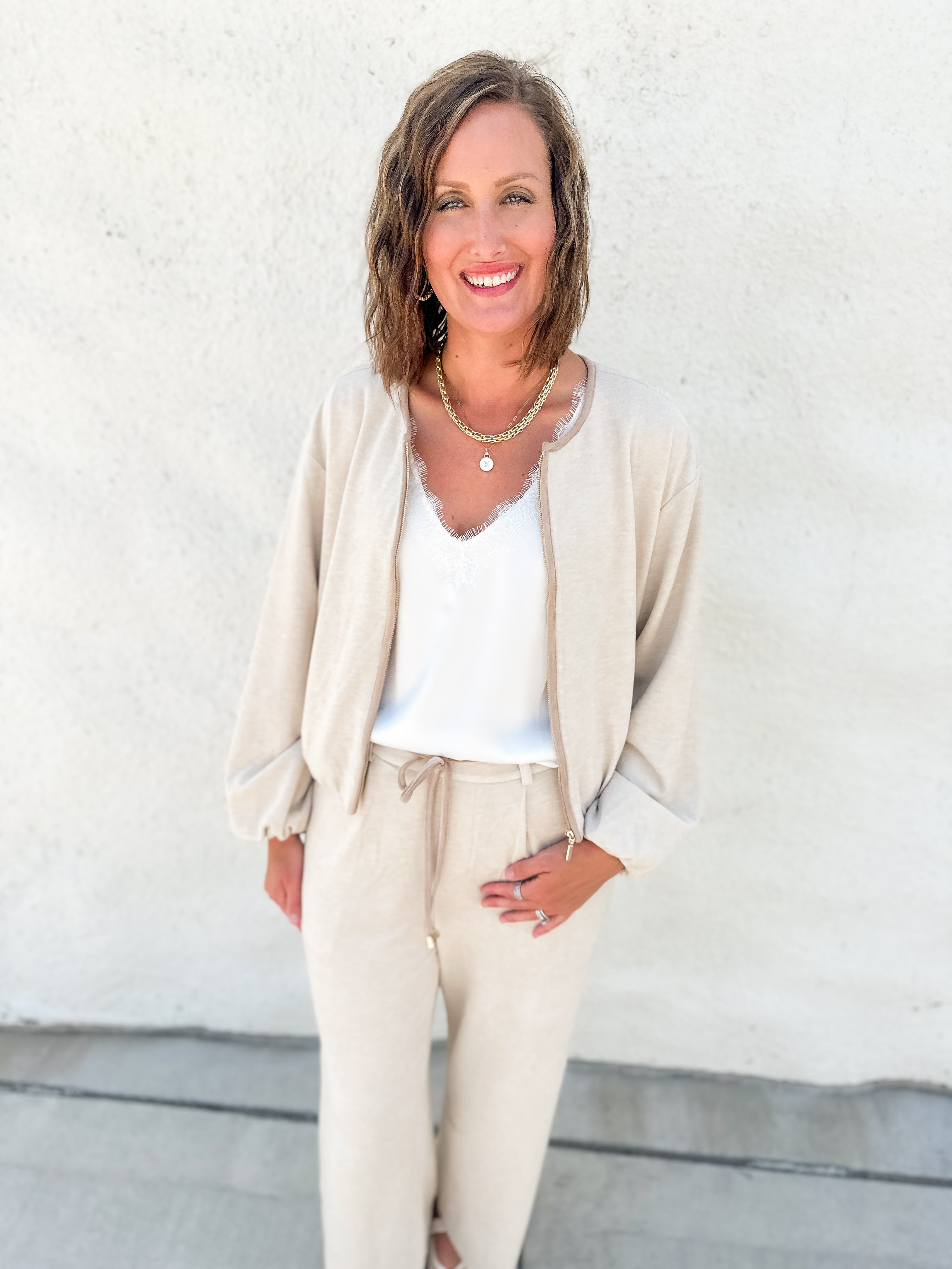 A woman smiling and posing in a beige bubble hem sweater bomber jacket with suede accents and matching drawstring pants over a white top, standing against a textured white stucco wall.