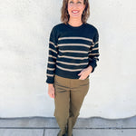 A woman stands outdoors against a white stucco wall wearing a black and beige horizontally striped knit sweater, olive green pants, and dark loafers.