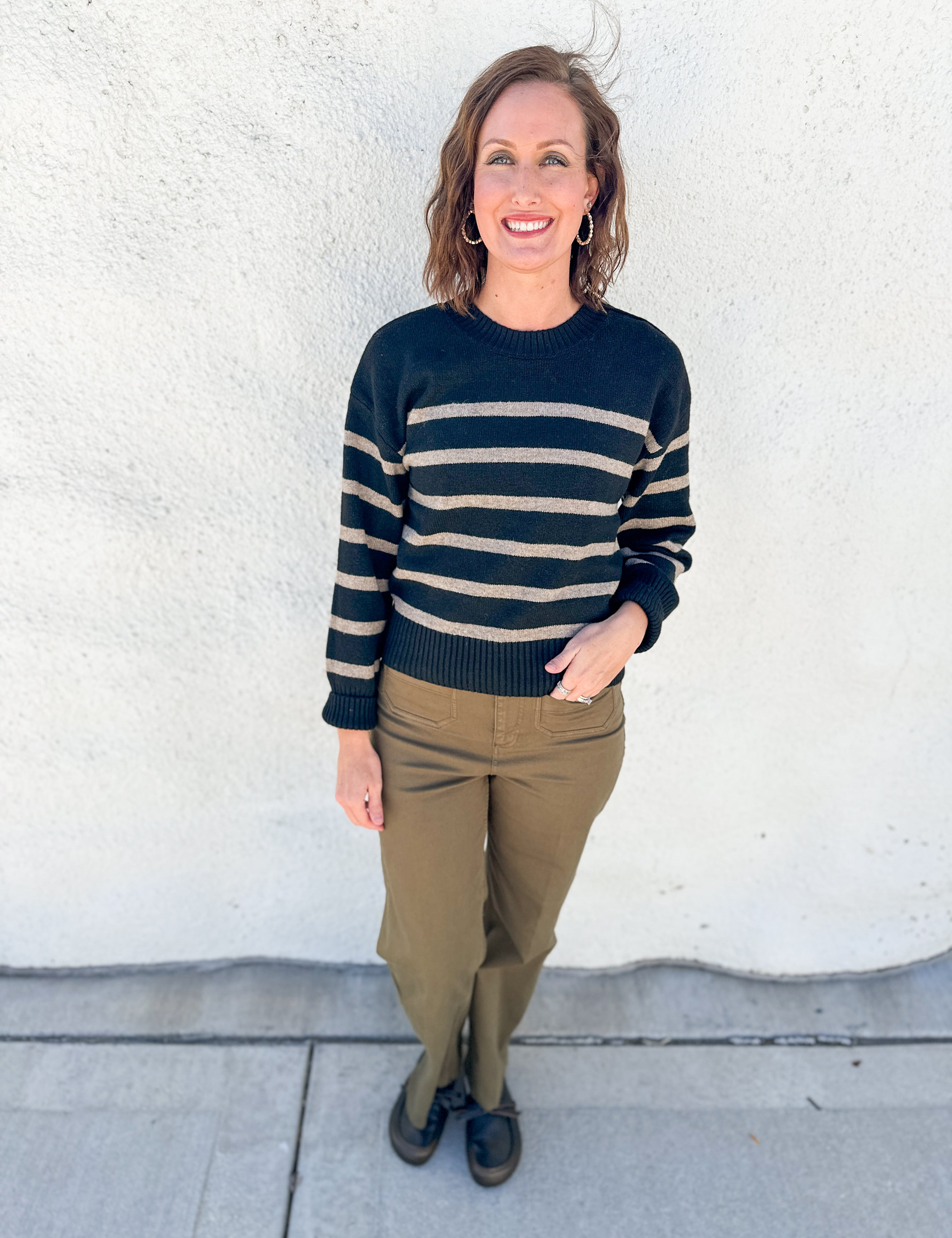 A woman stands outdoors against a white stucco wall wearing a black and beige horizontally striped knit sweater, olive green pants, and dark loafers.