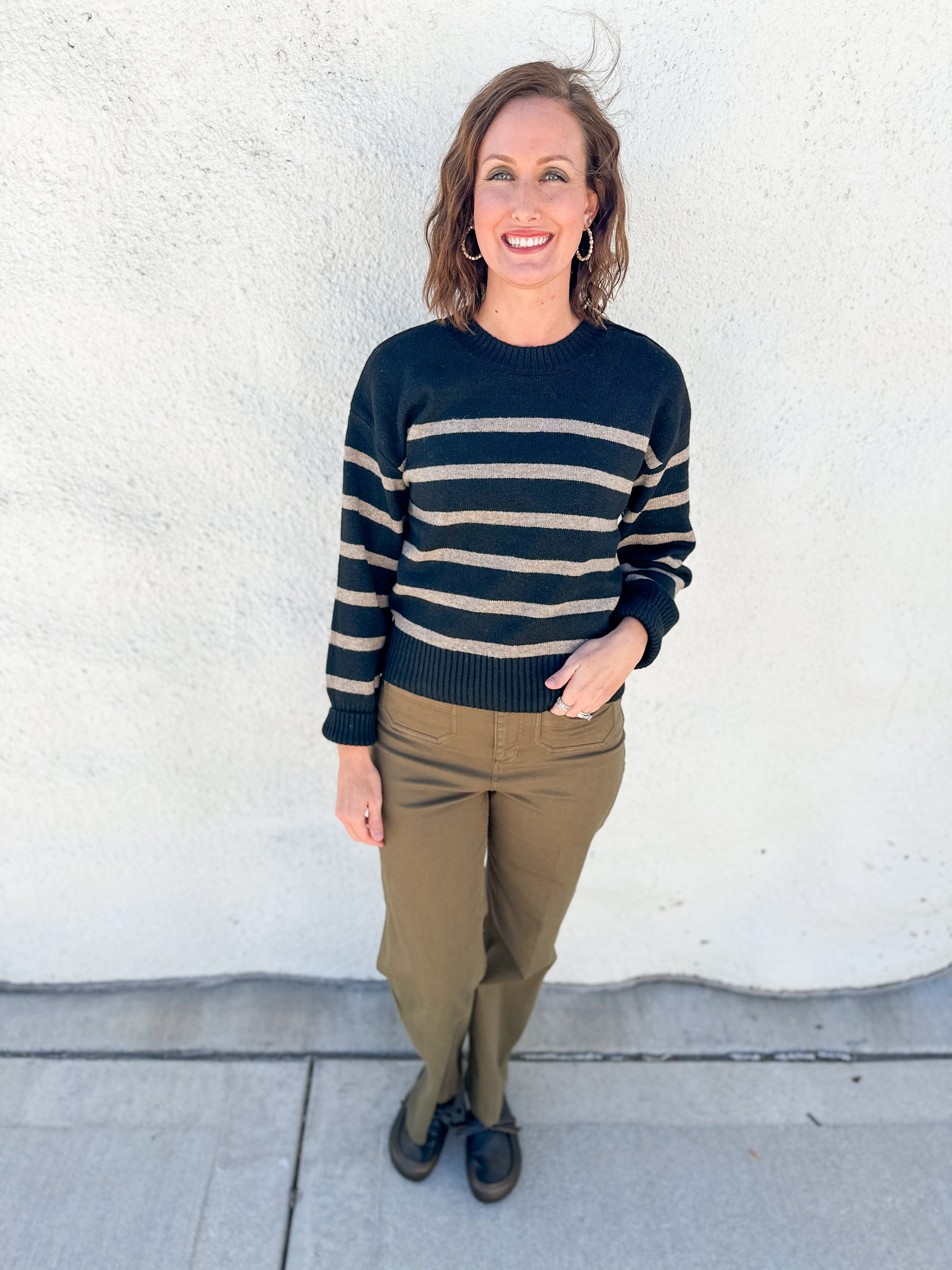 A woman stands outdoors against a white stucco wall wearing a black and beige horizontally striped knit sweater, olive green pants, and dark loafers.
