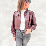 A woman wearing a cropped, shiny burgundy faux-leather jacket with puffed sleeves, styled over a white top and gray jeans, standing against a textured white wall.
