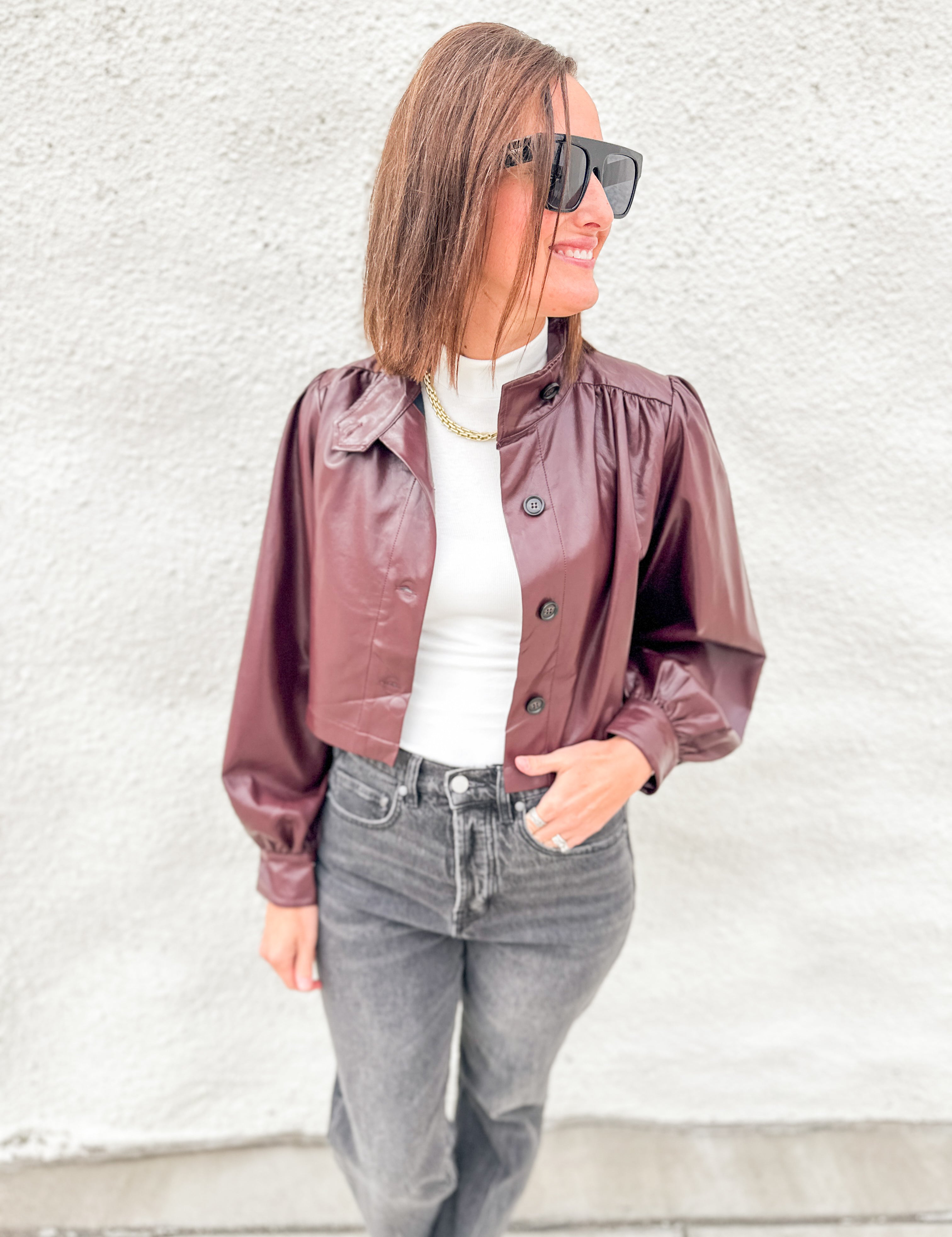 A woman wearing a cropped, shiny burgundy faux-leather jacket with puffed sleeves, styled over a white top and gray jeans, standing against a textured white wall.