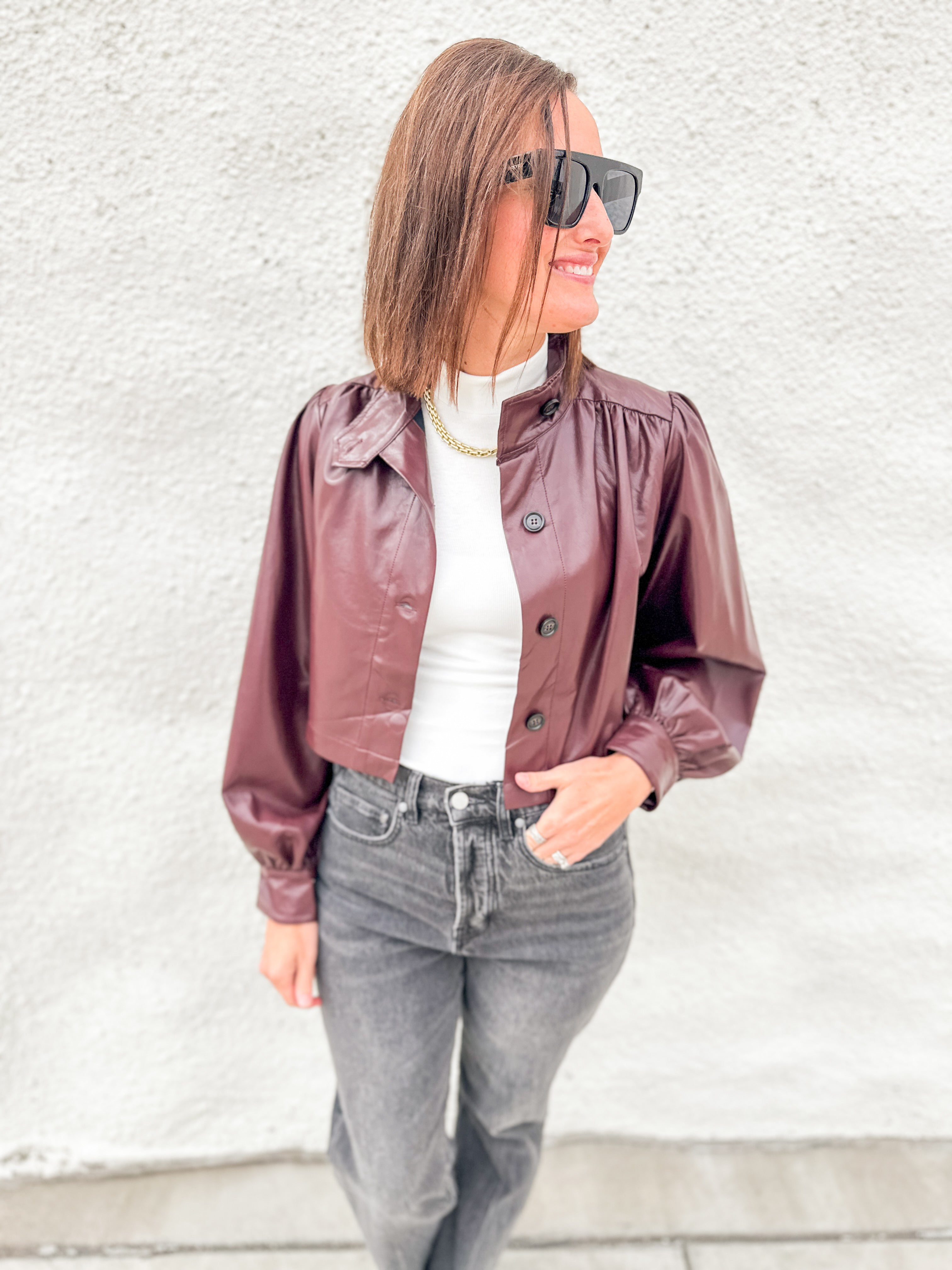 A woman wearing a cropped, shiny burgundy faux-leather jacket with puffed sleeves, styled over a white top and gray jeans, standing against a textured white wall.