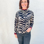 A woman with short brown hair smiling, wearing a black and beige zebra-print mock neck sweater with dark gray jeans, standing against a white textured wall.