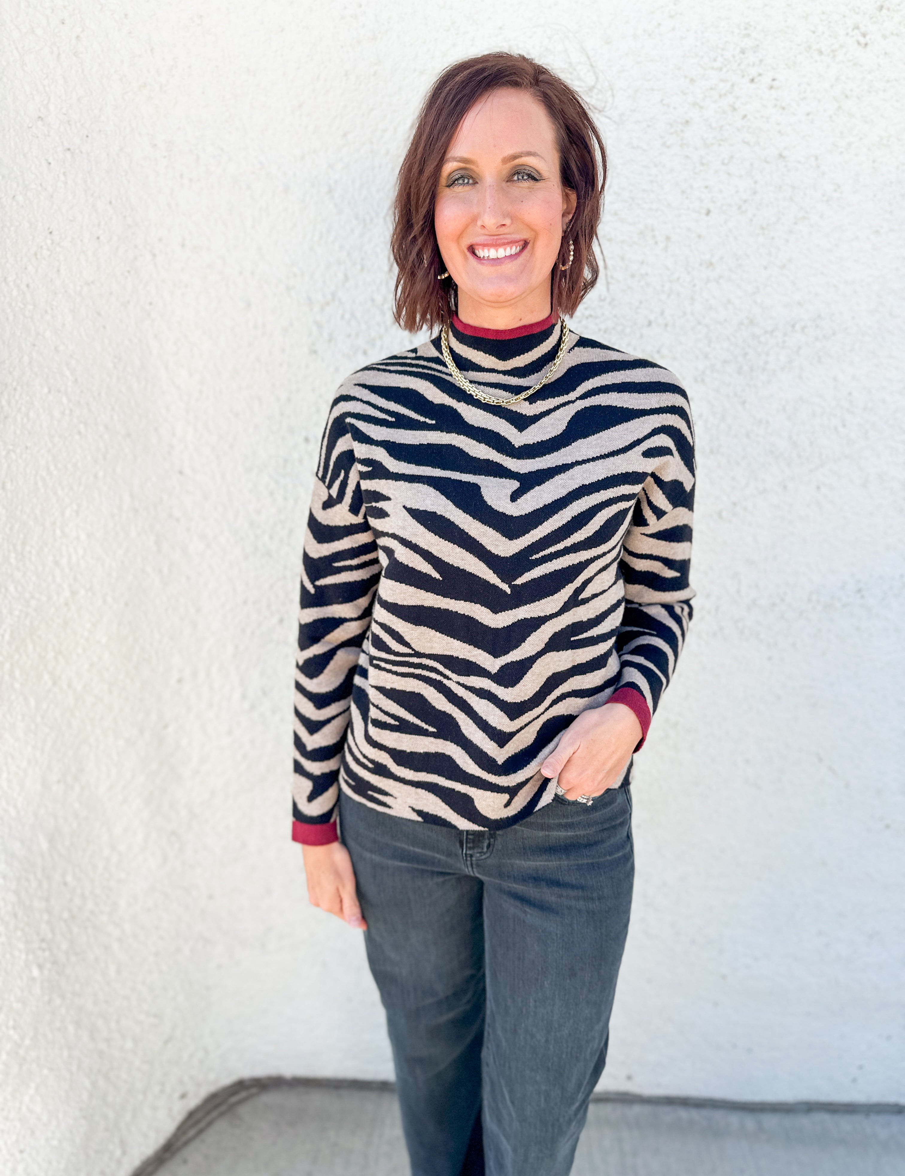 A woman with short brown hair smiling, wearing a black and beige zebra-print mock neck sweater with dark gray jeans, standing against a white textured wall.