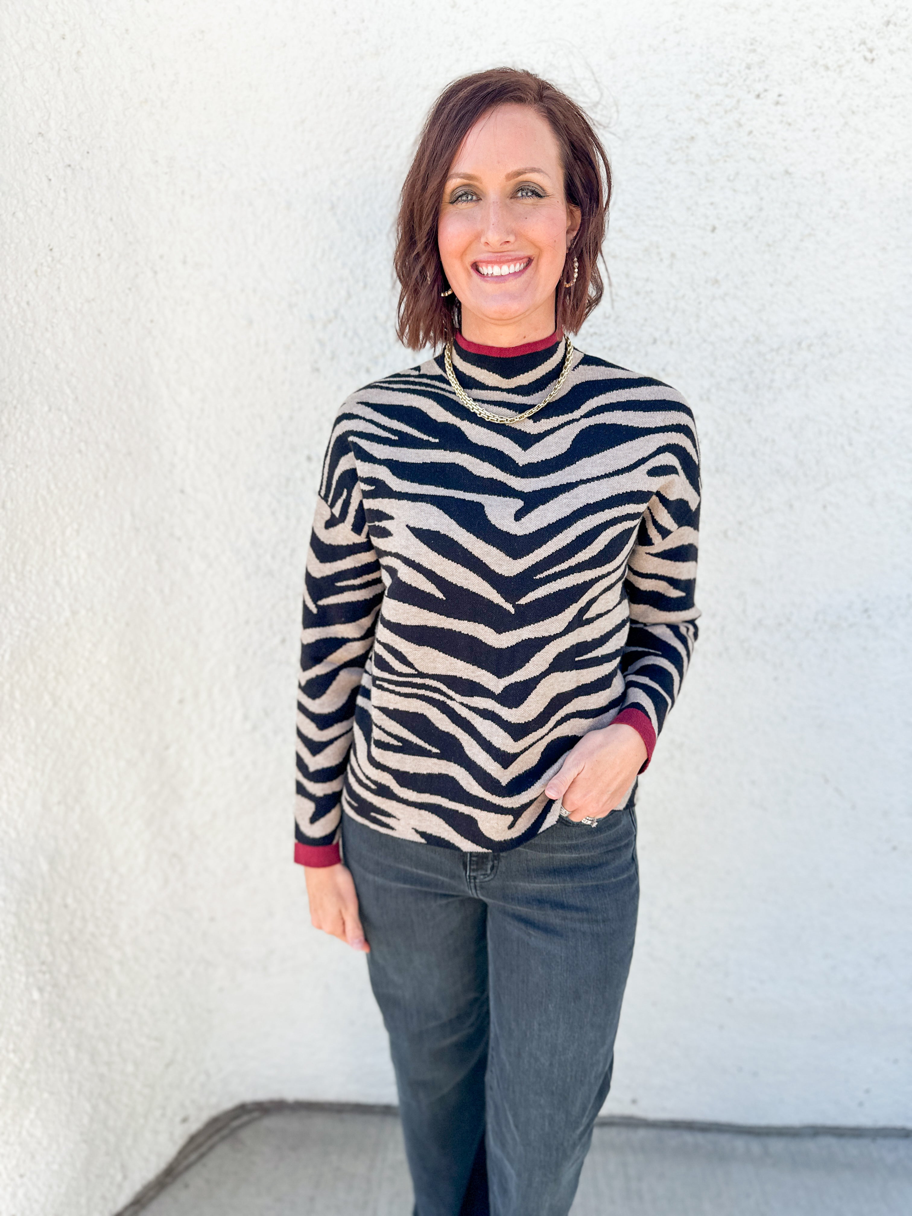 A woman with short brown hair smiling, wearing a black and beige zebra-print mock neck sweater with dark gray jeans, standing against a white textured wall.