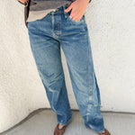 A person wearing blue wide-leg barrel denim jeans, a gray and white striped long-sleeve shirt, and brown flats, standing against a textured off-white wall.