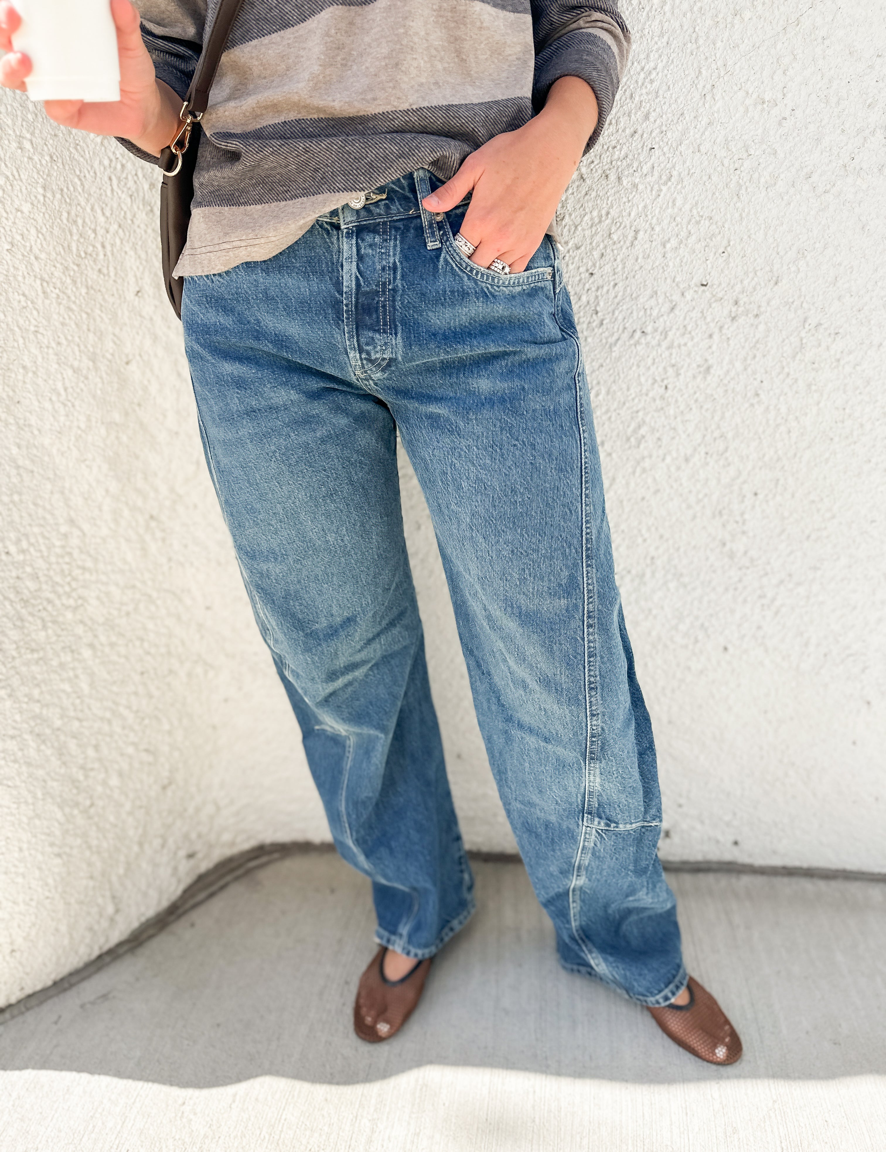 A person wearing blue wide-leg barrel denim jeans, a gray and white striped long-sleeve shirt, and brown flats, standing against a textured off-white wall.