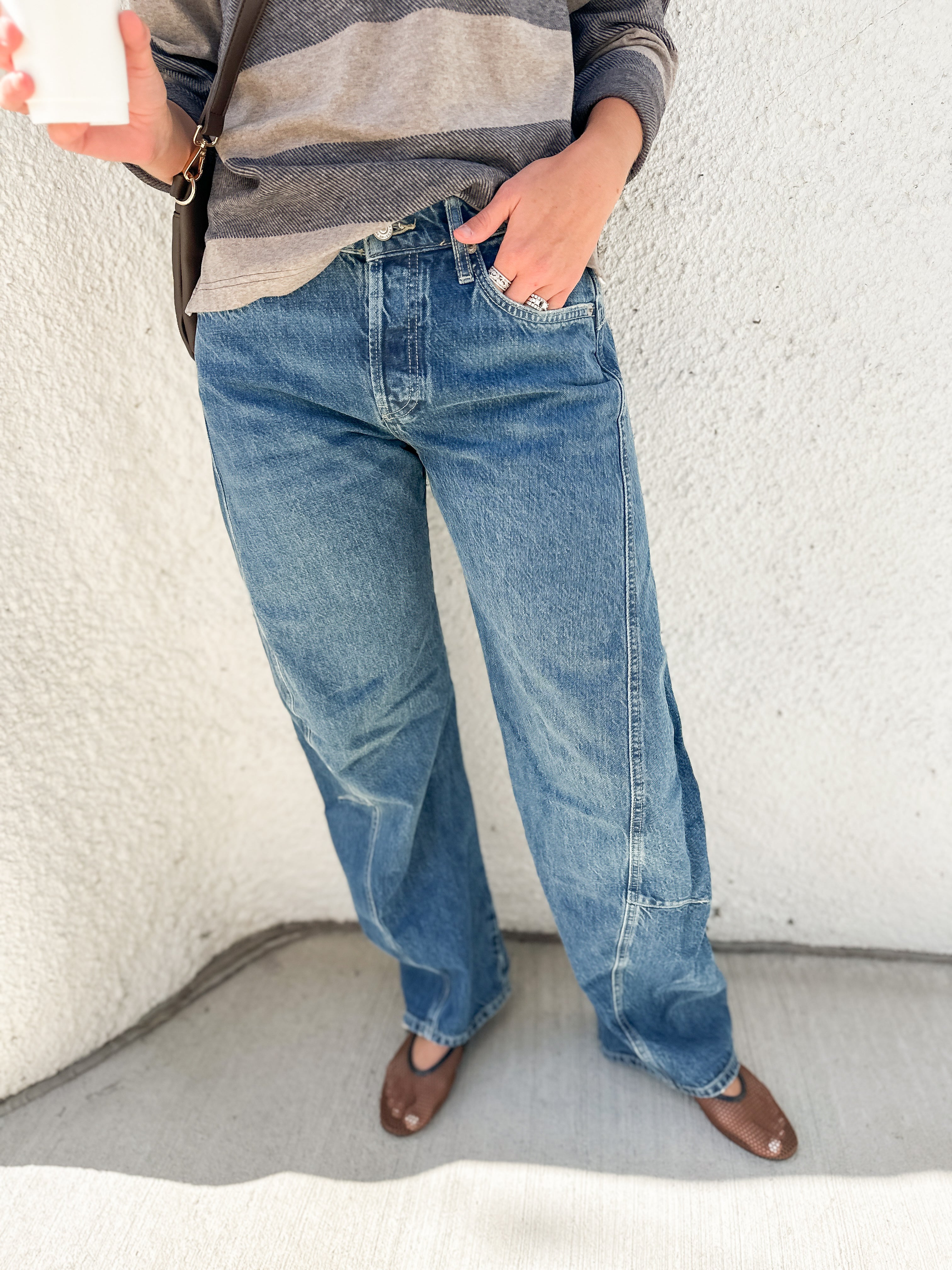 A person wearing blue wide-leg barrel denim jeans, a gray and white striped long-sleeve shirt, and brown flats, standing against a textured off-white wall.