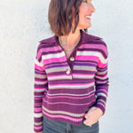 A woman wearing a purple, pink, and gray striped knit sweater by Free People with a buttoned henley collar, paired with dark gray jeans, standing against a light gray textured wall.