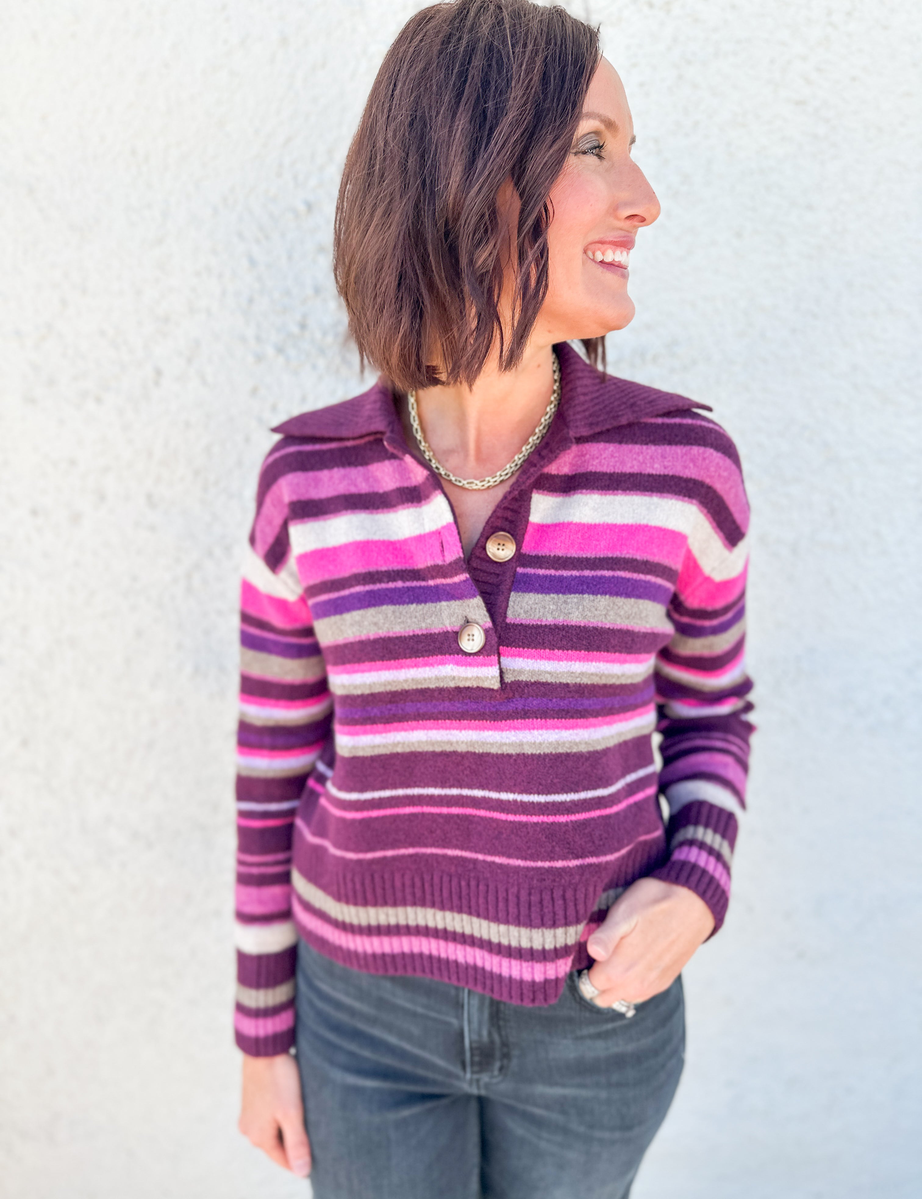A woman wearing a purple, pink, and gray striped knit sweater by Free People with a buttoned henley collar, paired with dark gray jeans, standing against a light gray textured wall.