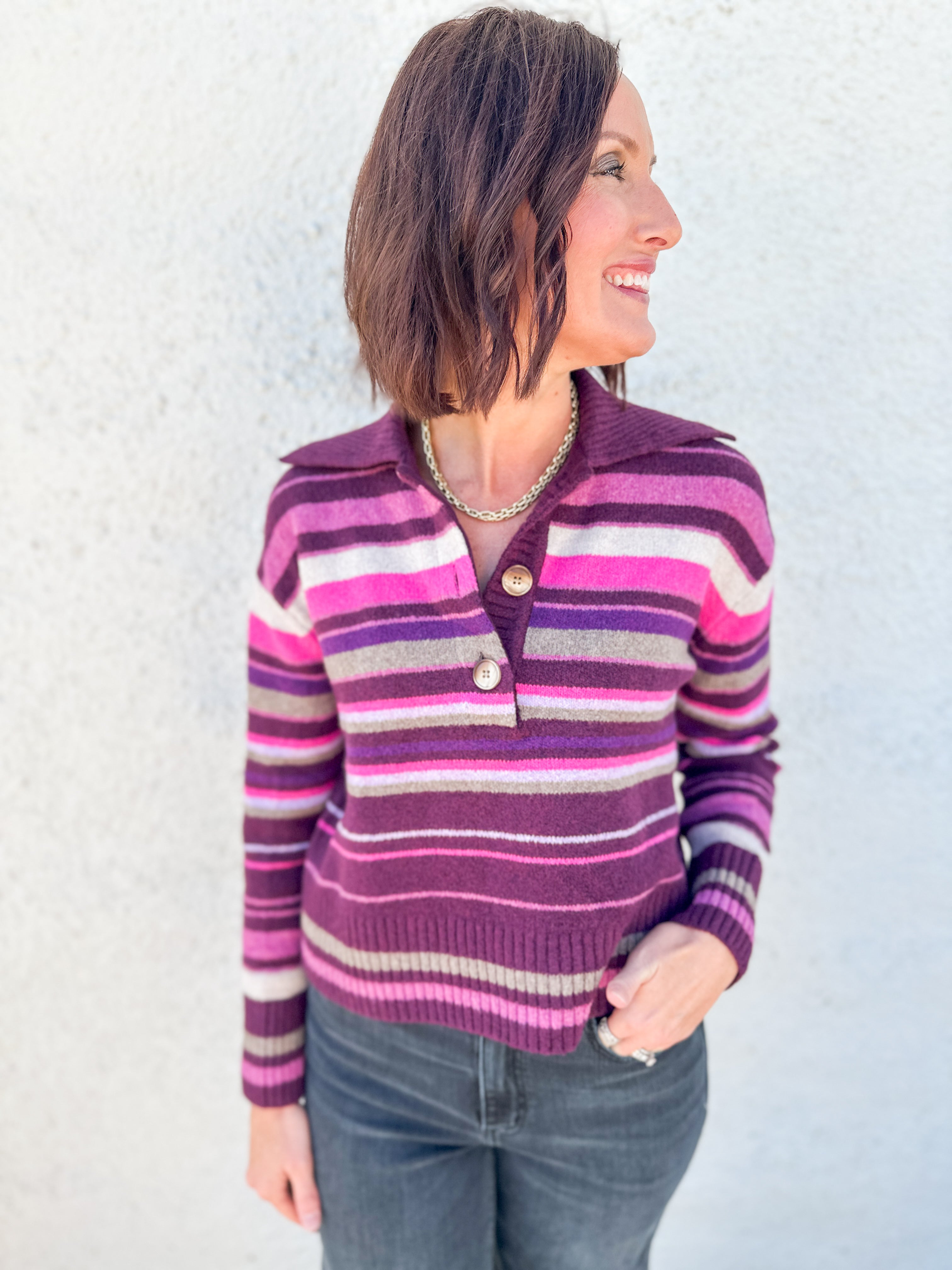 A woman wearing a purple, pink, and gray striped knit sweater by Free People with a buttoned henley collar, paired with dark gray jeans, standing against a light gray textured wall.
