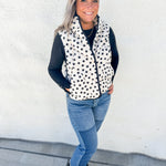 A woman wearing a black and white polka dot zip-up vest over a black long-sleeve top, paired with blue jeans and black pointed-toe flats, standing against a white stucco wall.