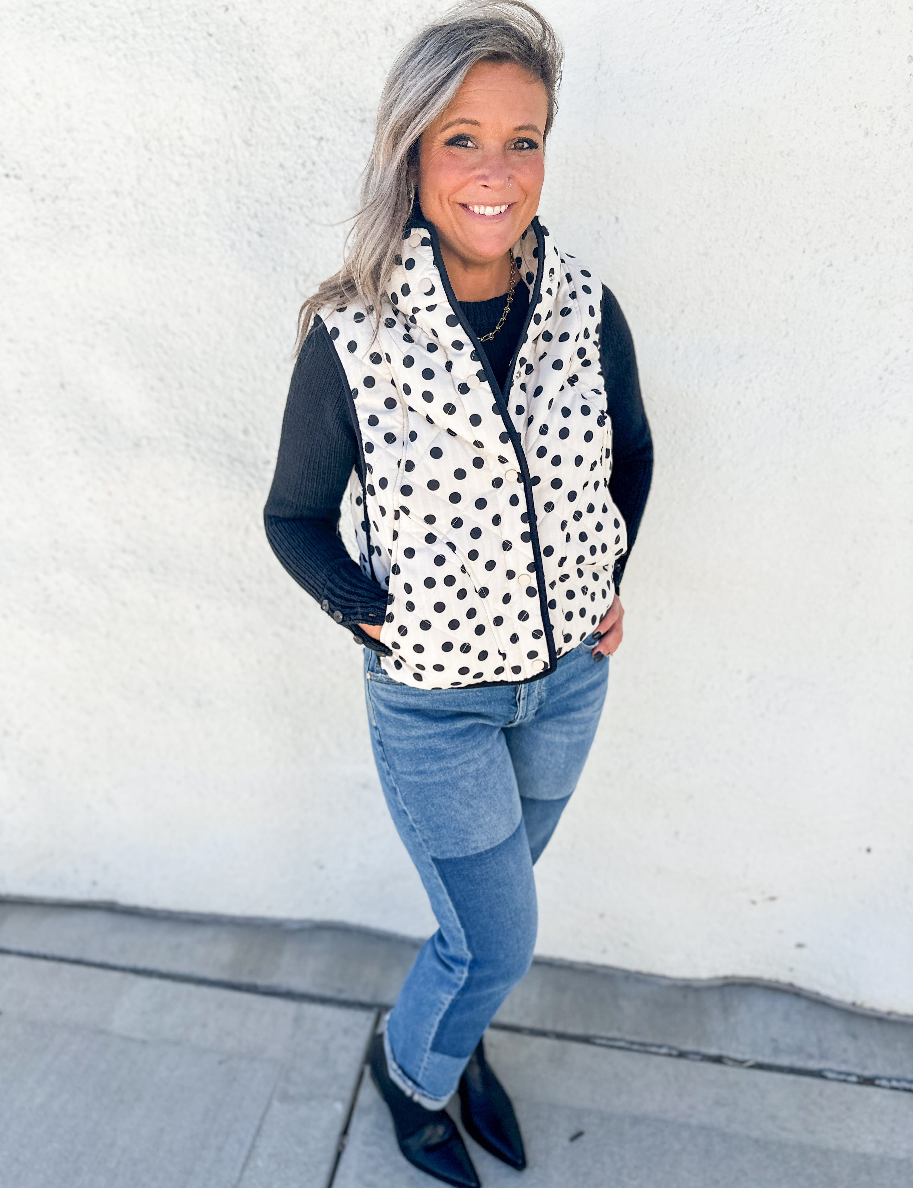 A woman wearing a black and white polka dot zip-up vest over a black long-sleeve top, paired with blue jeans and black pointed-toe flats, standing against a white stucco wall.