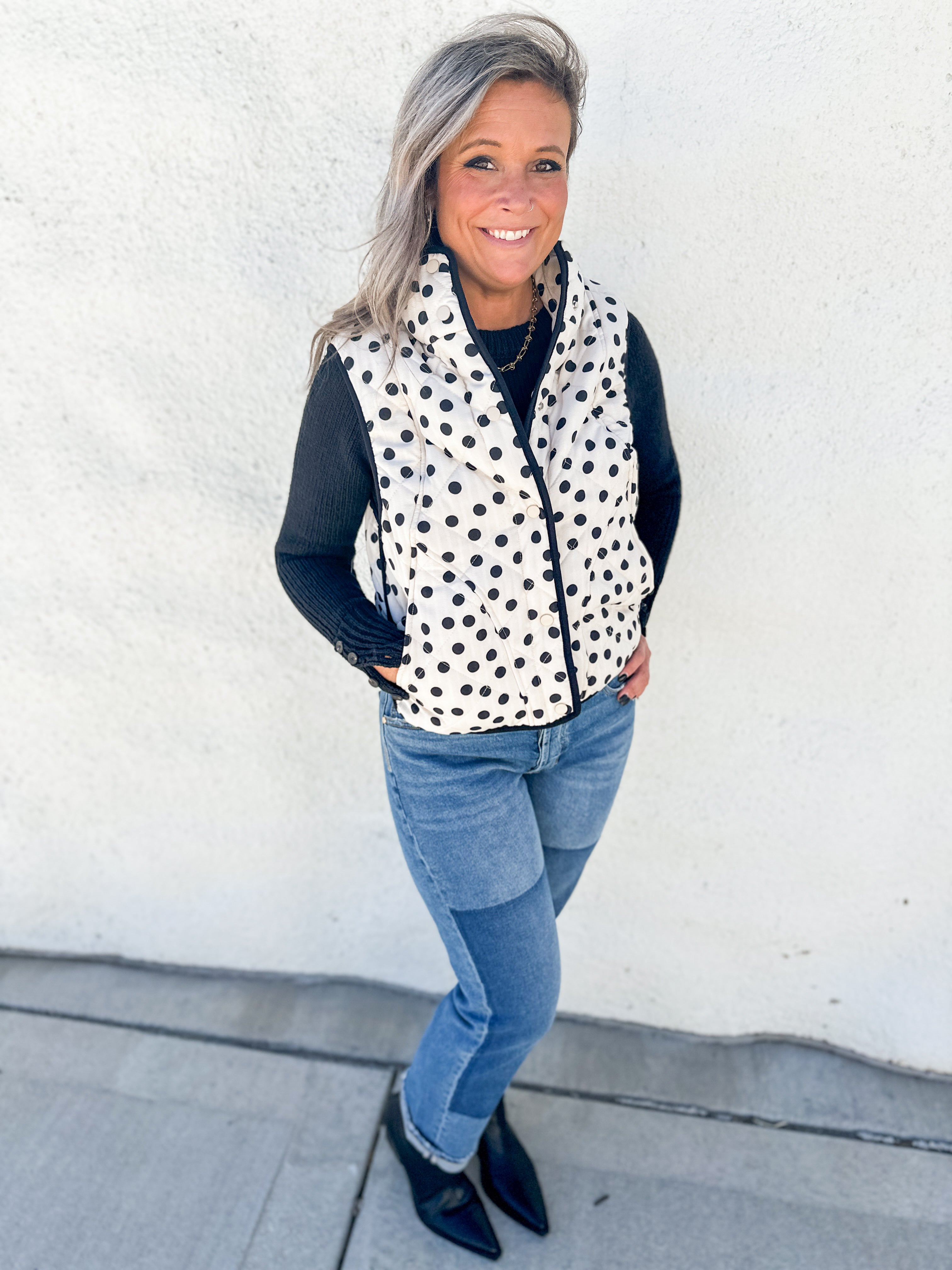 A woman wearing a black and white polka dot zip-up vest over a black long-sleeve top, paired with blue jeans and black pointed-toe flats, standing against a white stucco wall.