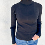 A woman wearing a black ribbed mock neck long-sleeve sweater and light blue jeans, standing against a textured white wall.